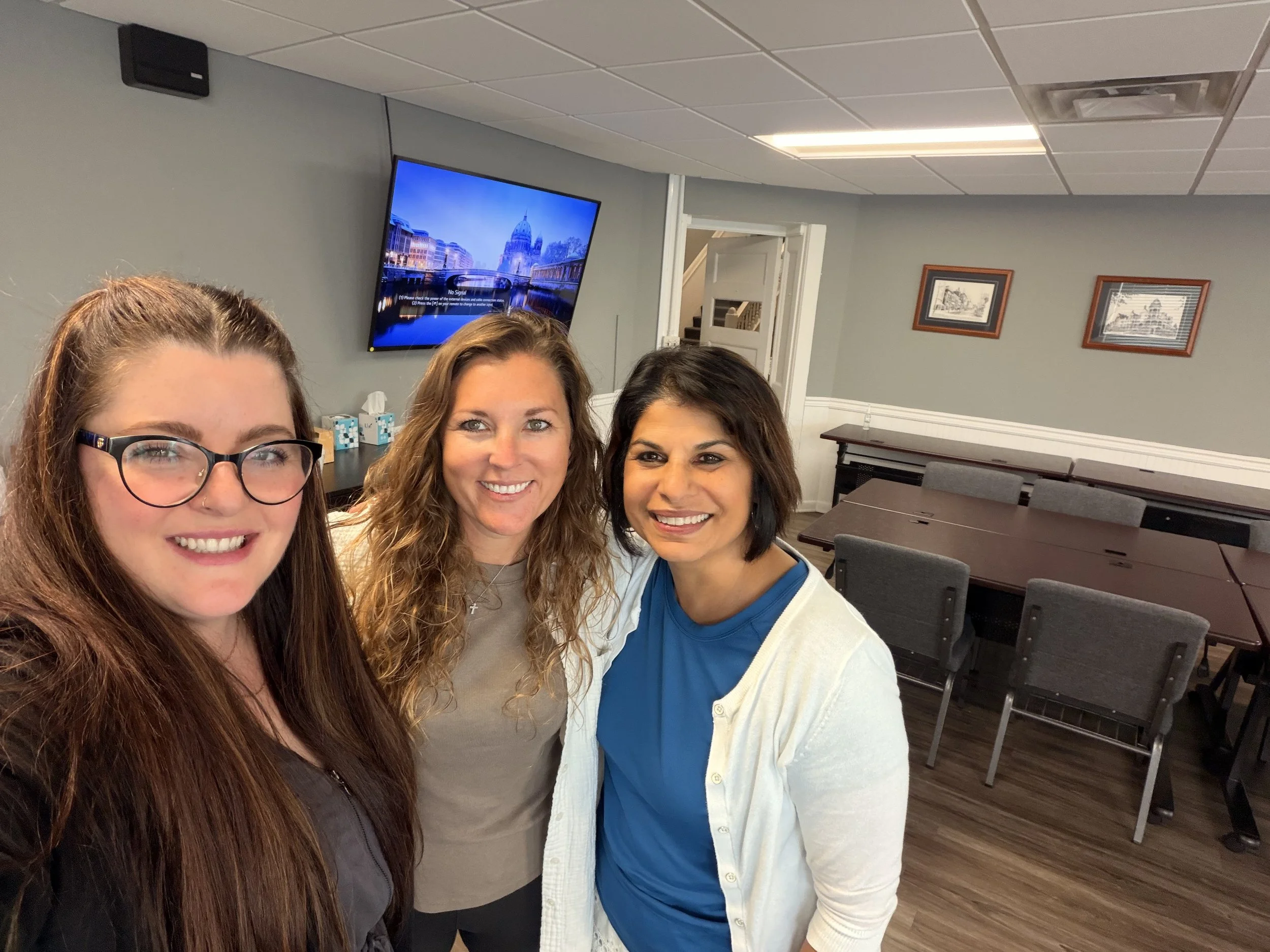 Four women smiling for a selfie in a room with gray walls, framed pictures, and a television displaying a cityscape with a river and bridge.