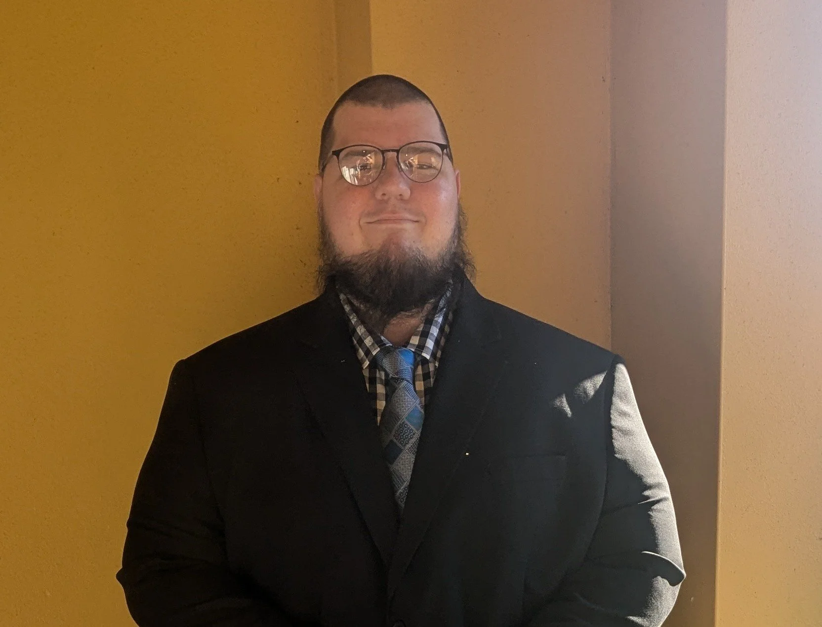 A man wearing glasses, a black suit, a checkered shirt, and a tie, standing against a two-toned beige and yellow wall.