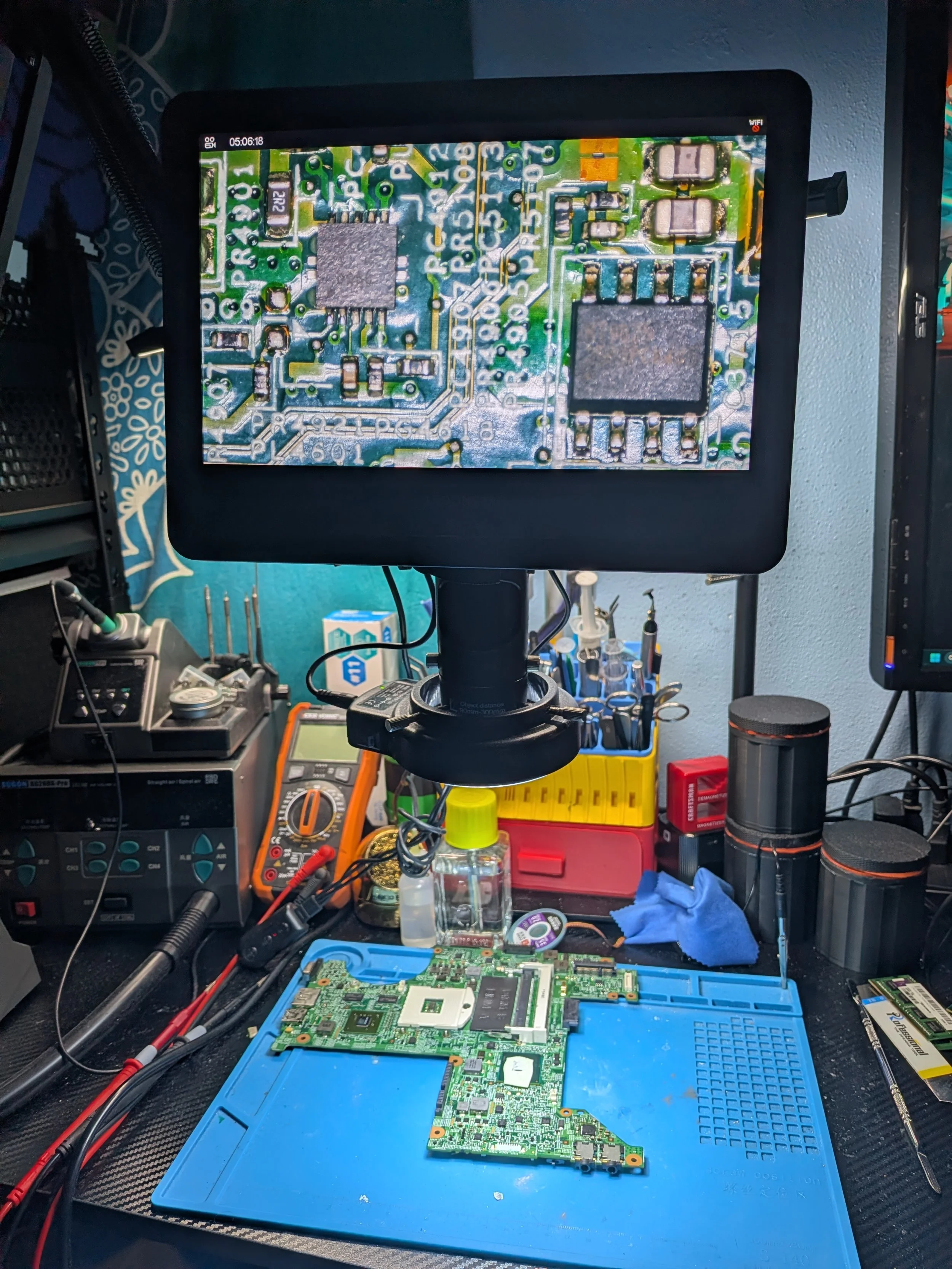 Close-up of a computer repair workspace with a circuit board on a blue anti-static mat, tools, and electronic equipment surrounding it.