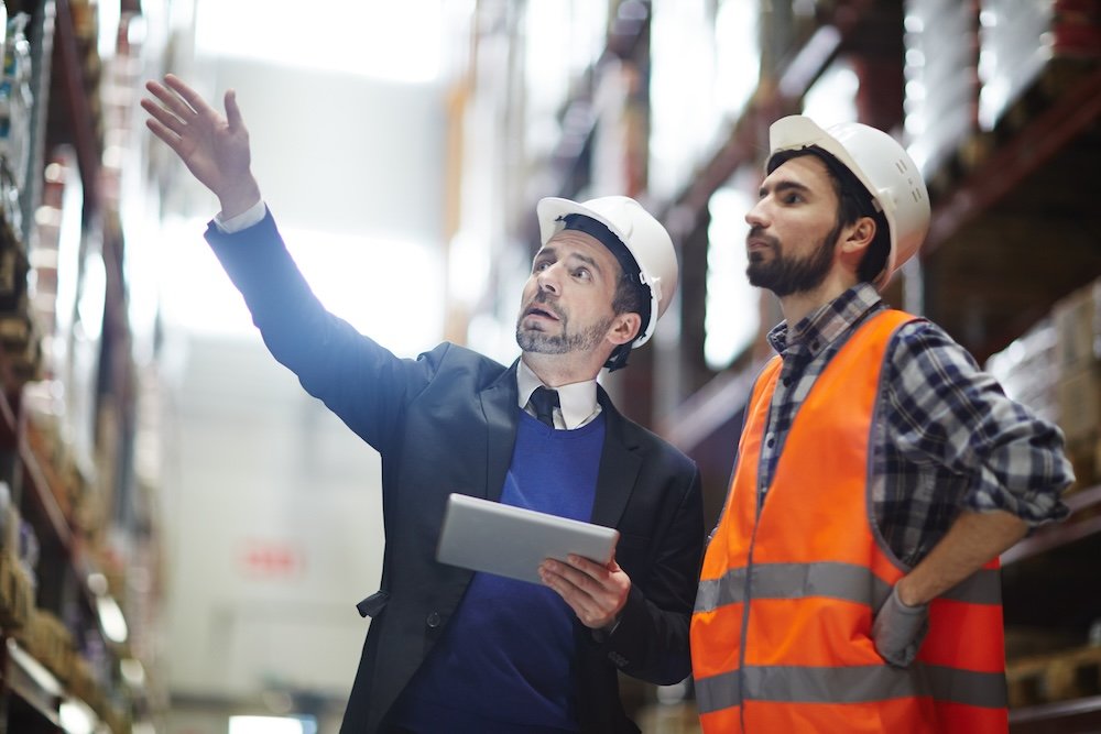 Two workers in hard hats and safety gear observing warehouse shelves