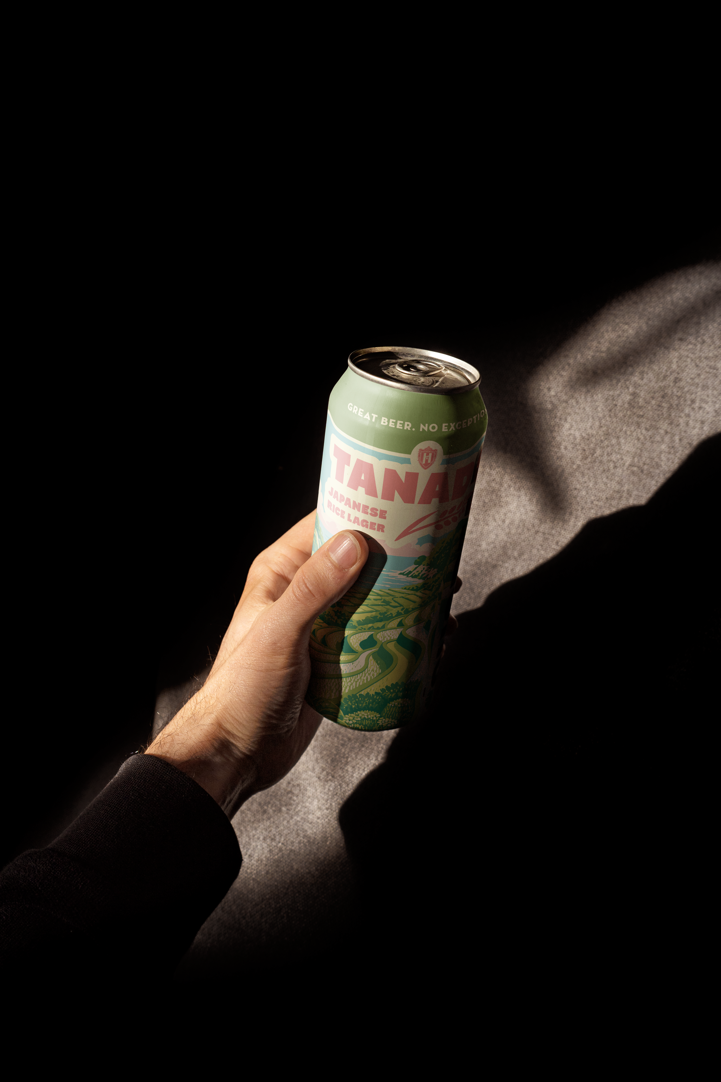 Close-up of a person's hand holding a can of Tanuki Japanese rice lager with a black background and a patterned gray surface.