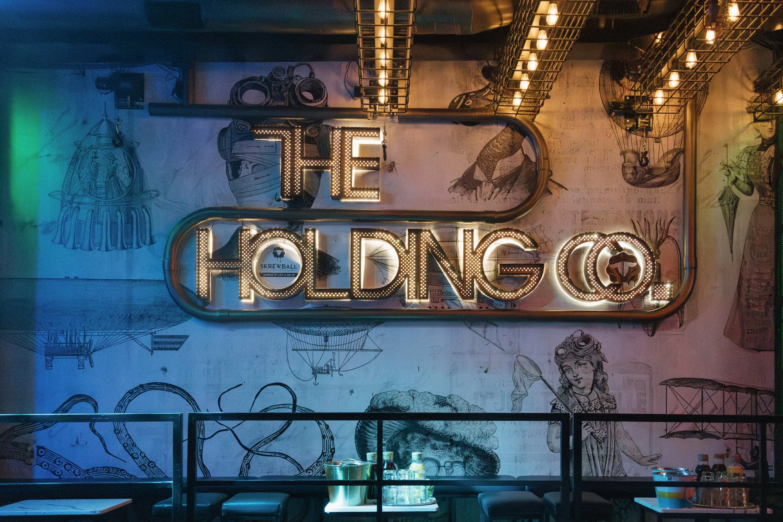 A dimly lit indoor space with wall art and a marquee sign spelling 'THE POD' in bright lights. The wall features sketches and drawings of various objects and figures, creating an artistic backdrop.
