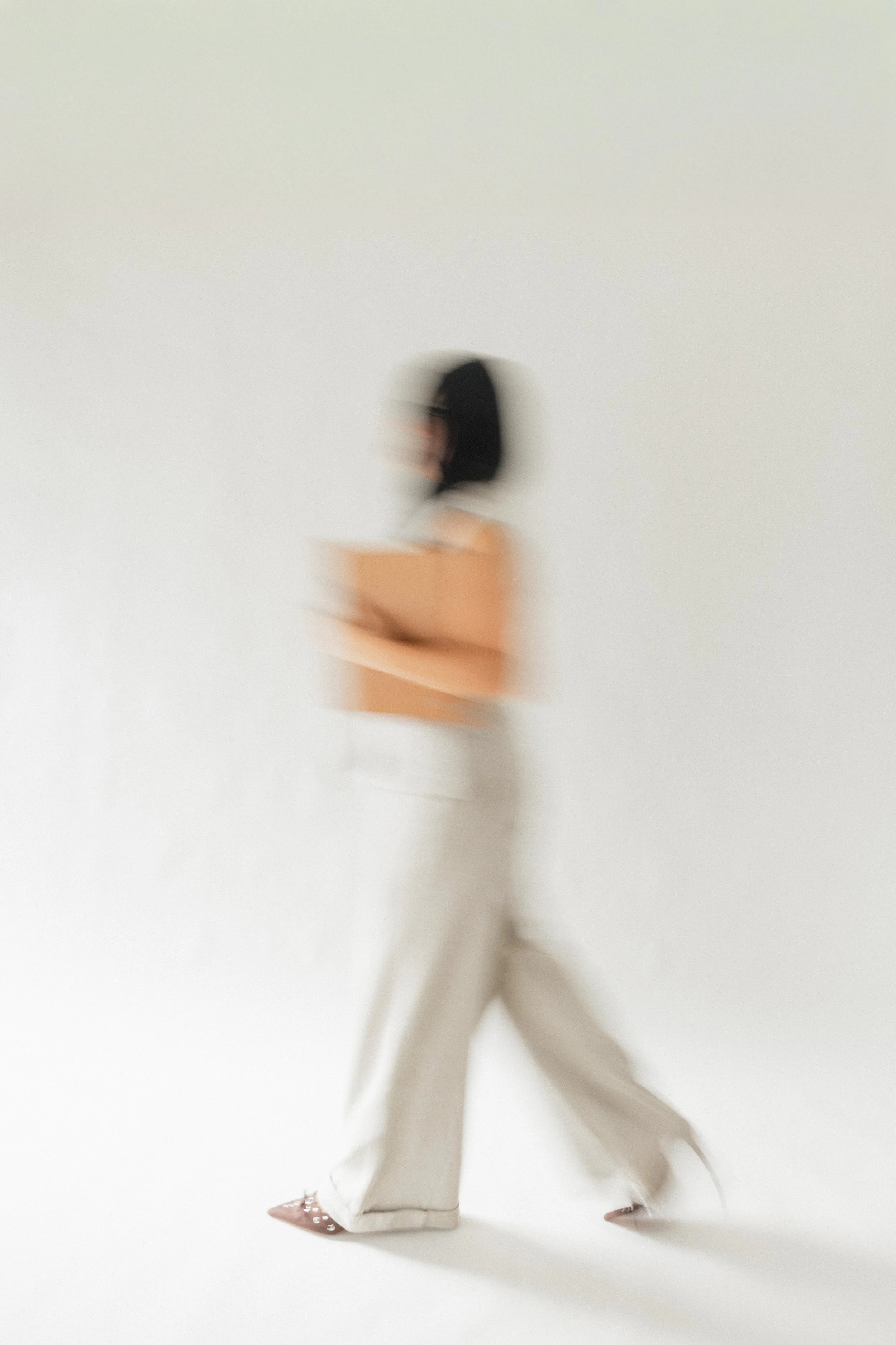 A blurred woman with black hair, wearing a white shirt, beige pants, and beige high heels, standing against a plain white background.