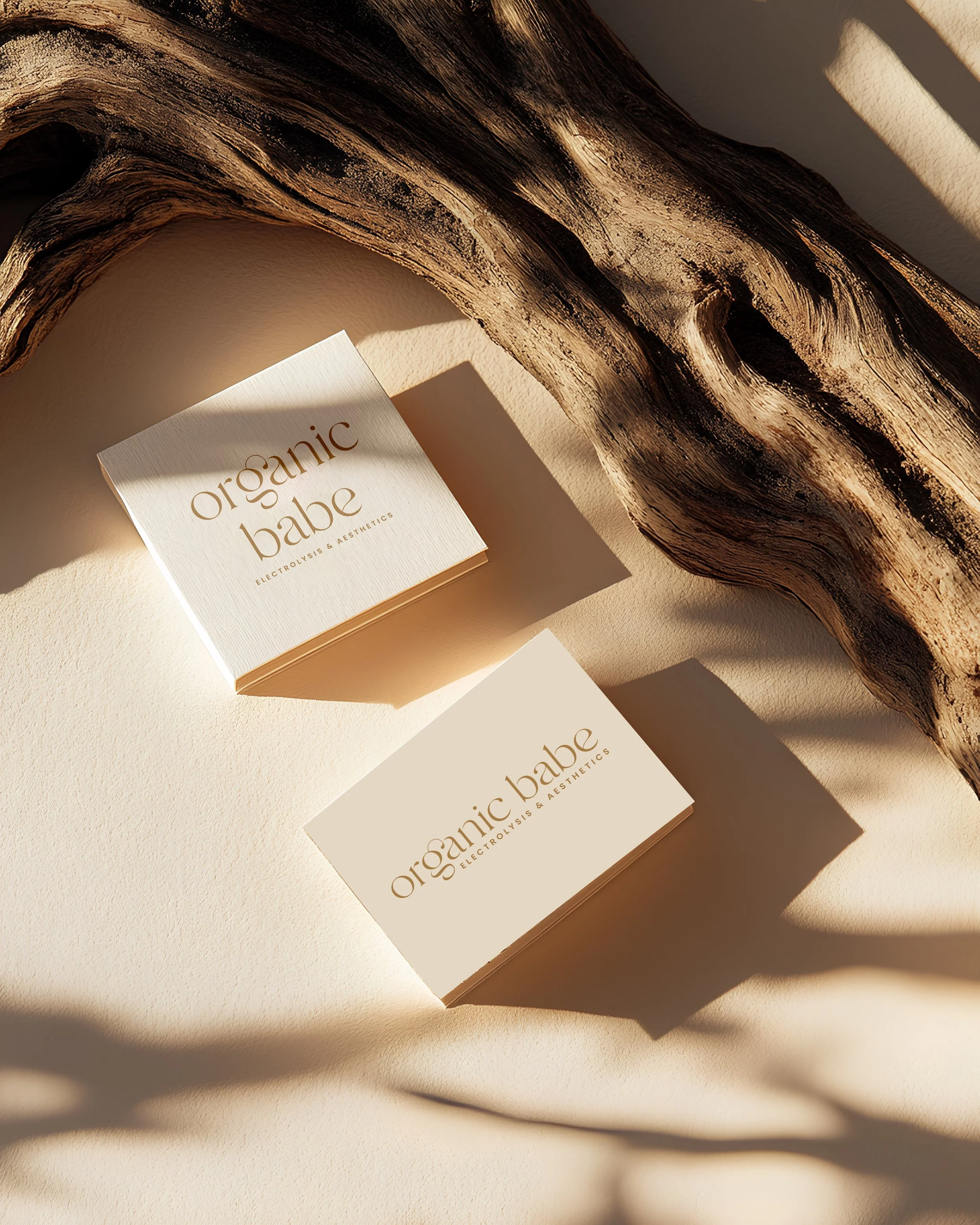 A clothing tag on a white surface with shadows. The tag reads "organic babe Electrolysis & Aesthetics" in gold lettering. A large piece of textured, dark painted wood is positioned above the tag, casting shadows on the surface.