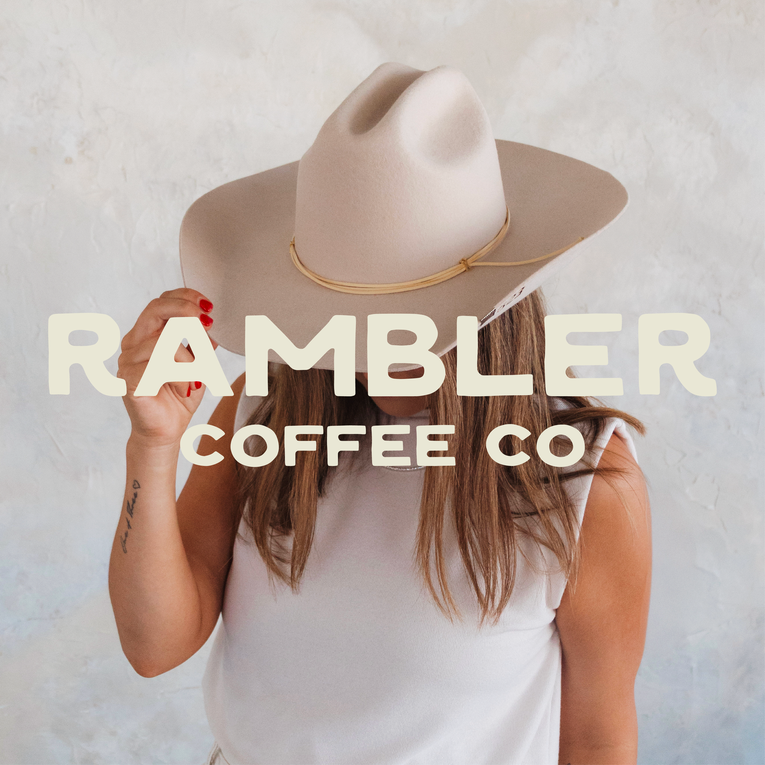 A woman with long hair wearing a white sleeveless top is holding a cardboard sign with large white letters that read 'RAMBLER' and other text partially visible, wearing a beige wide-brimmed hat with a string around it, against a plain light-colored wall.