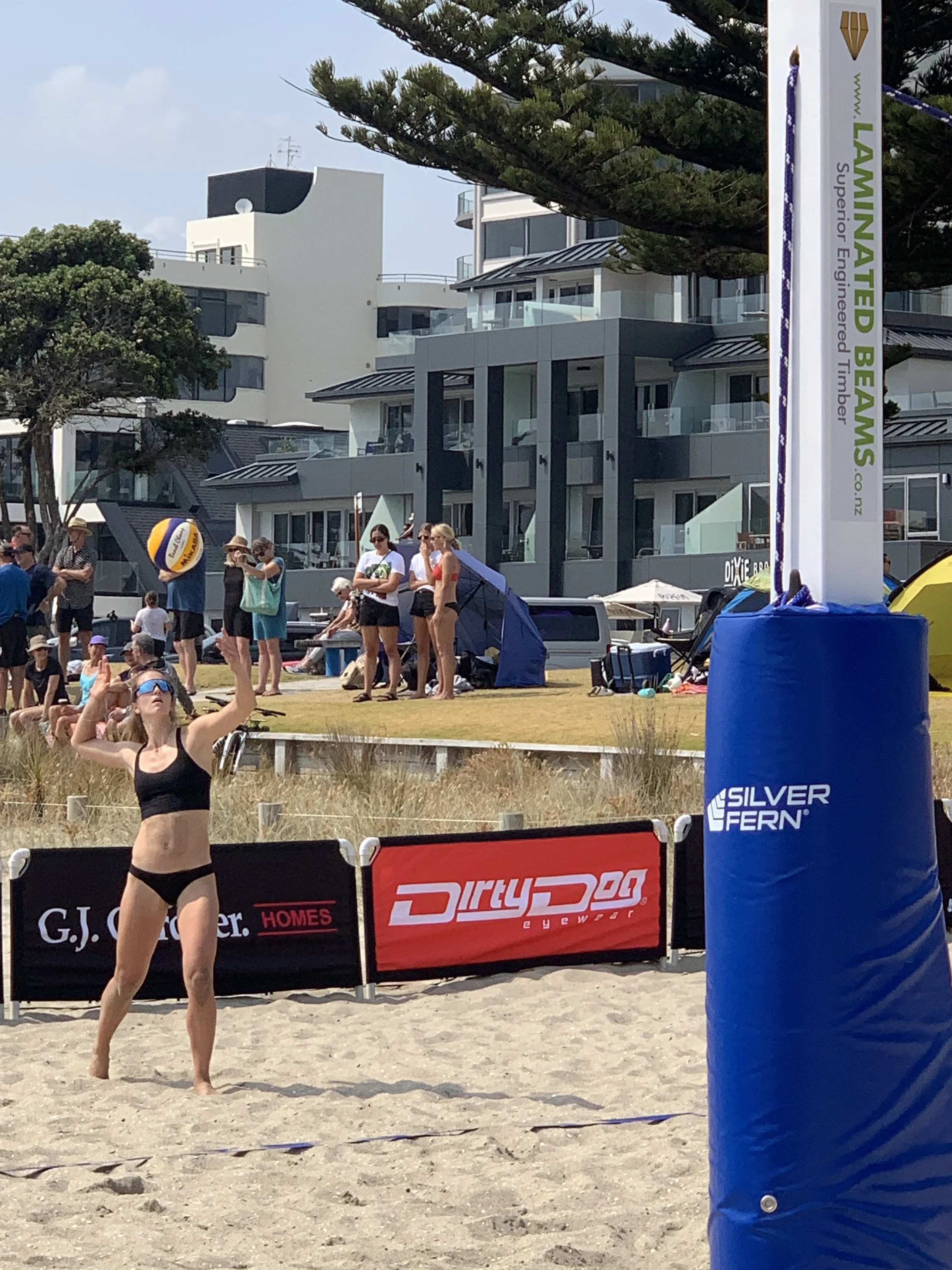 A woman in a black bikini playing beach volleyball on a sandy court, with a blue volleyball in the background and spectators watching behind a barrier. There are modern buildings and trees in the background, and a large blue padded pole in the foreground.