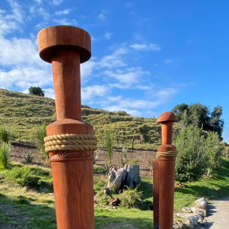 Two wooden posts wrapped with thick rope, outdoors in a grassy landscape with hills, trees, rocks, and a blue sky with clouds.