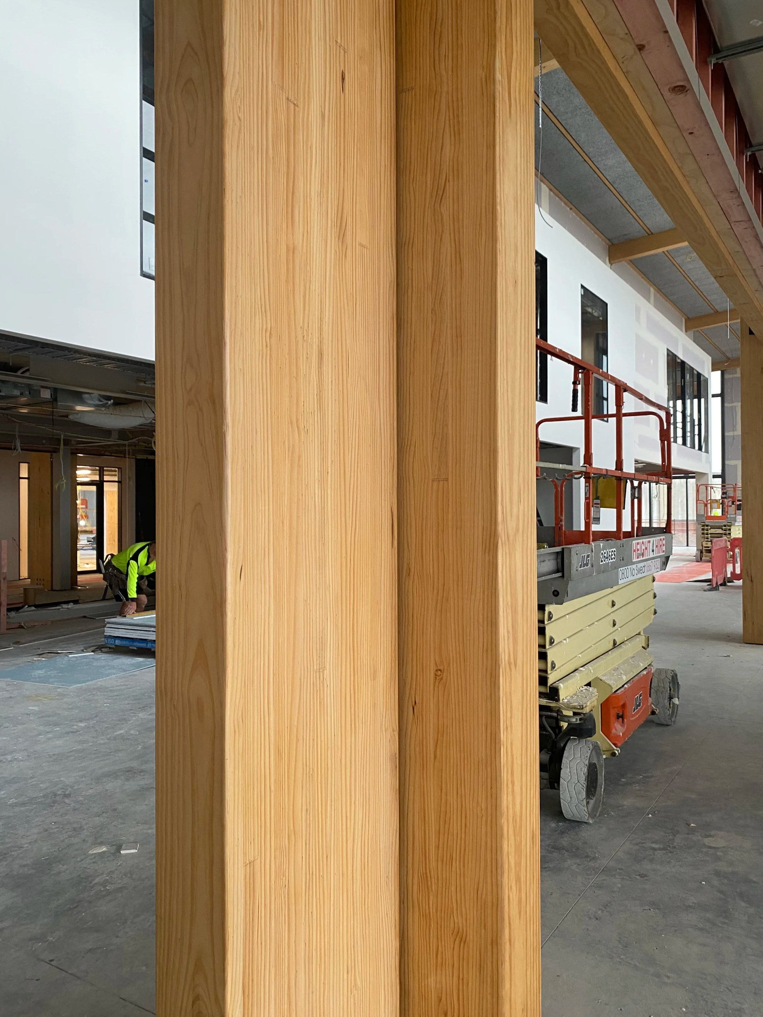 Interior of a building under construction with large wooden beams and a worker in a neon safety vest working on the floor.