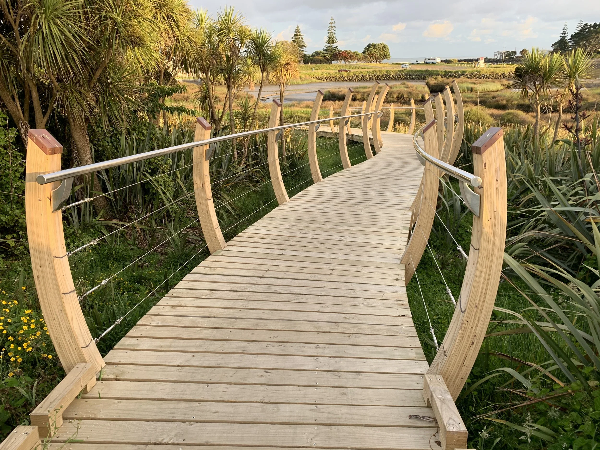 A wooden boardwalk with metal railings winding through lush green plants and trees, leading towards a marsh or wetland area with water, grass, and distant trees, under a partly cloudy sky.