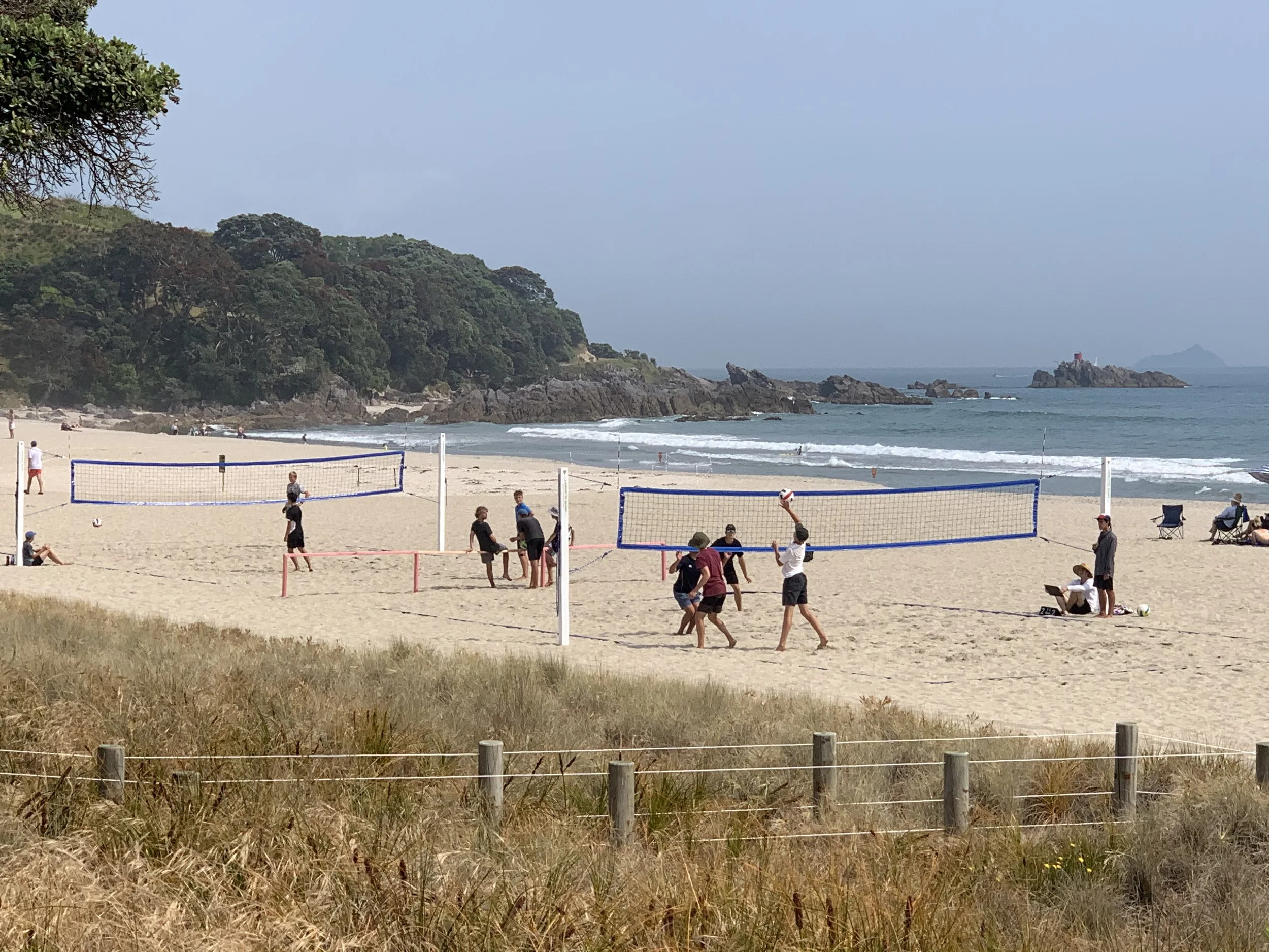 Beach scene with volleyball game, people playing and relaxing on the sand, rocky shoreline, ocean waves, and a lighthouse on a small rocky island in the distance.