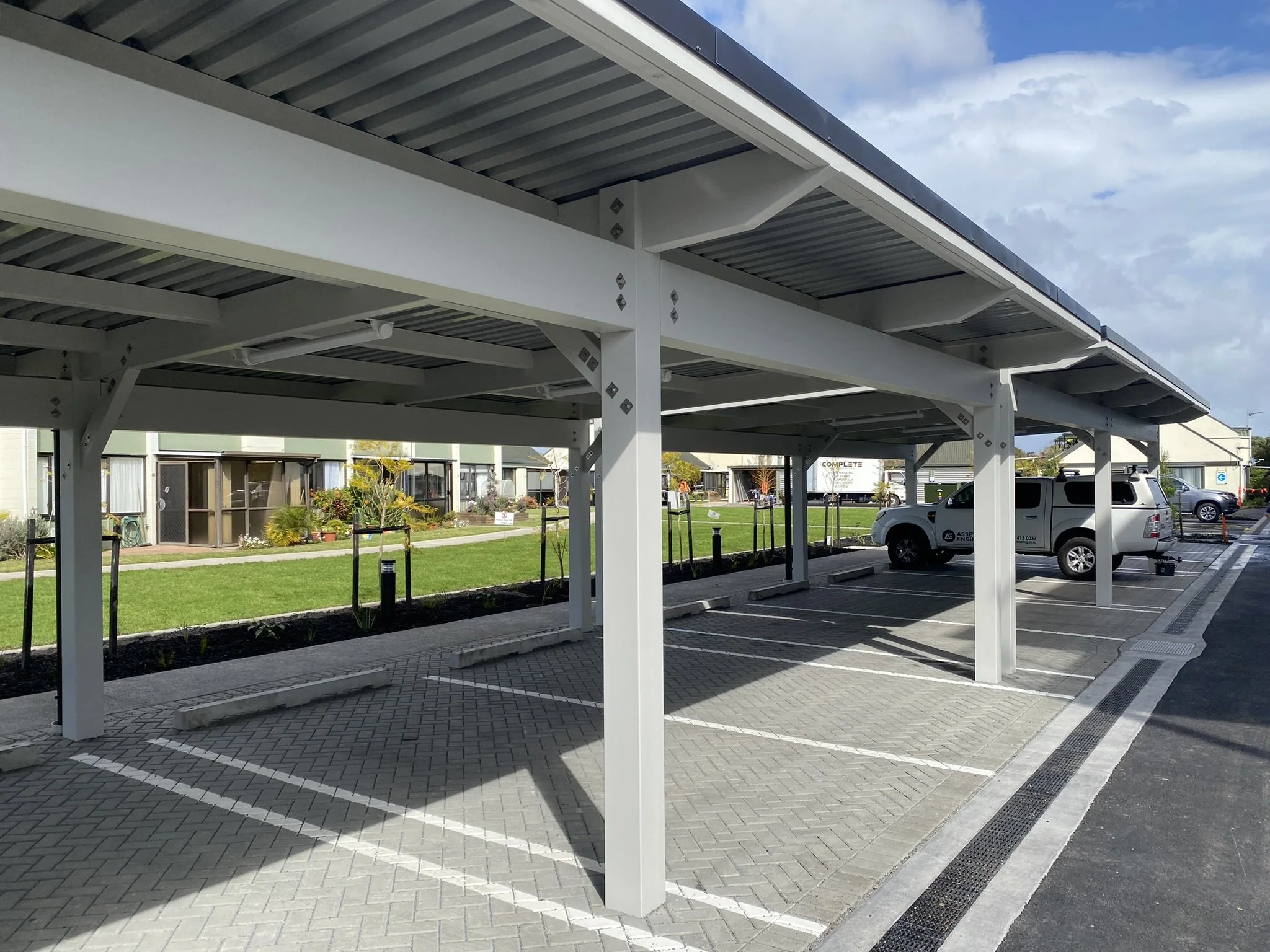 Empty parking area under a large metal shelter with a landscaped background and a cloudy sky.