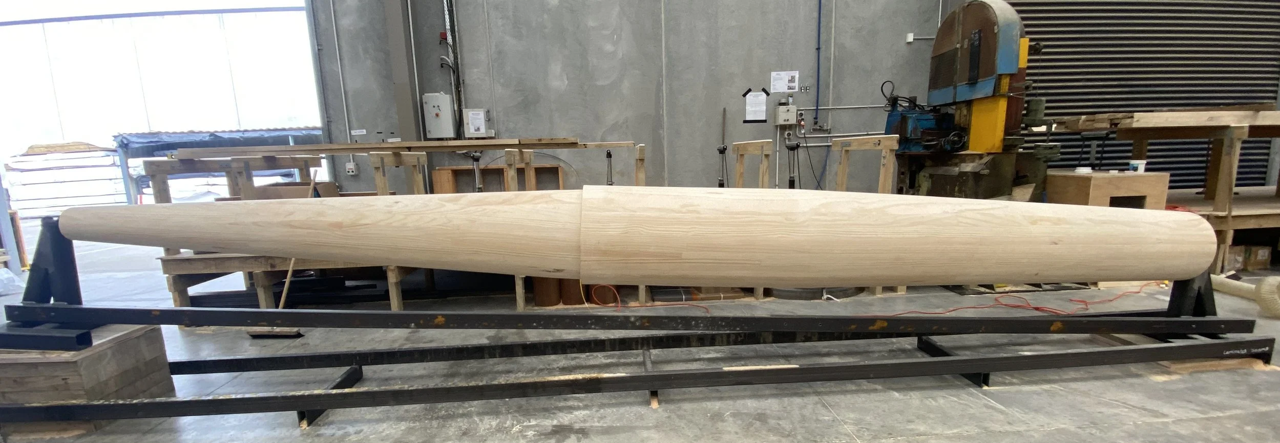 A large wooden kayak in a workshop, resting on a black frame with craftsmanship tools and equipment around.
