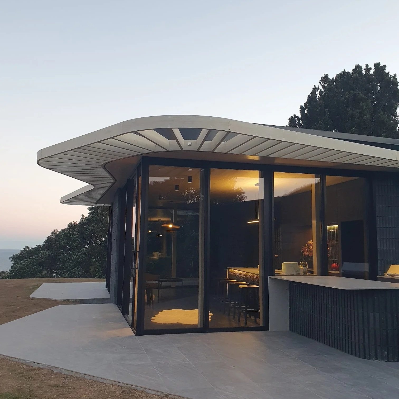 Modern house with large glass windows and a curved roof, situated outdoors during dusk, with trees in the background.