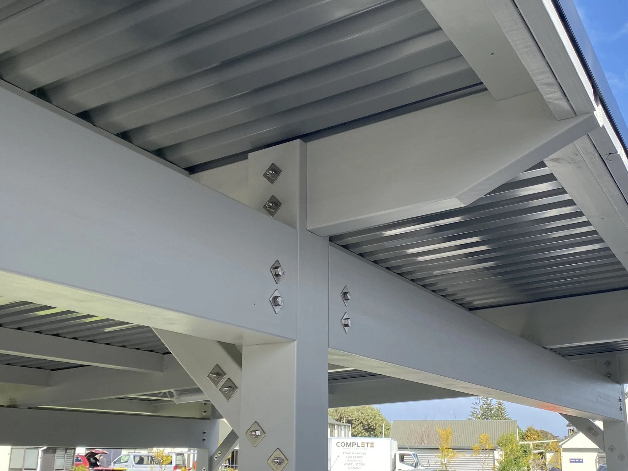 Close-up of a modern architectural structure showing metal beams and panels, with a background of blue sky and trees.