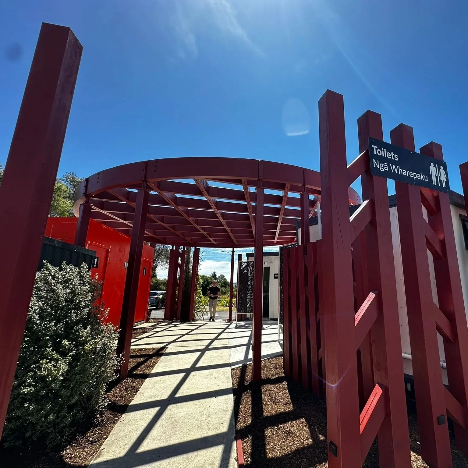 Outdoor entrance to public toilets marked with a sign, with red wooden fencing and a curved roof, under a bright sunny sky.