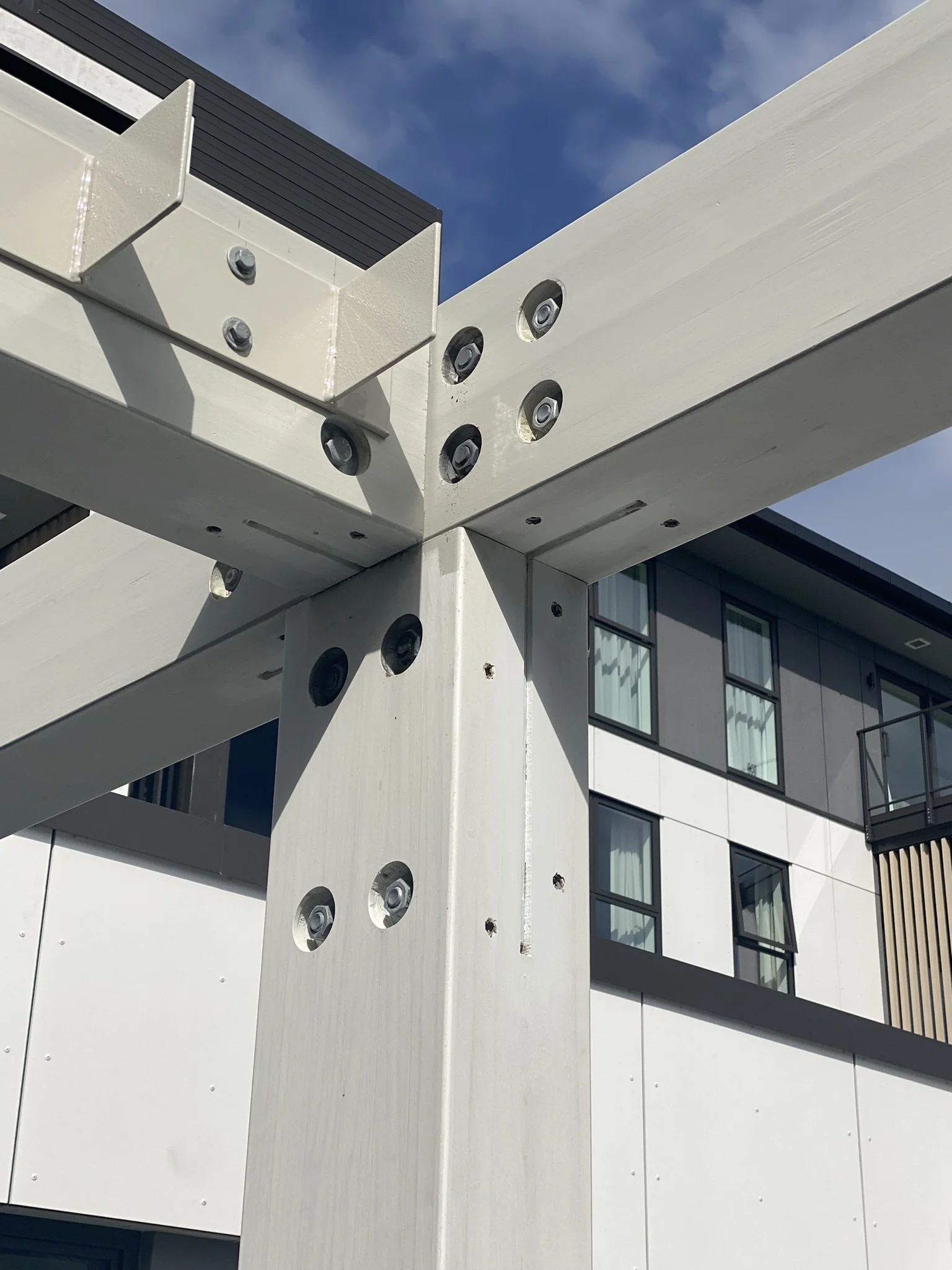 Close-up of a modern building's exterior showing white structural beams with embedded bolts, and glass windows with black frames, under a partly cloudy sky.