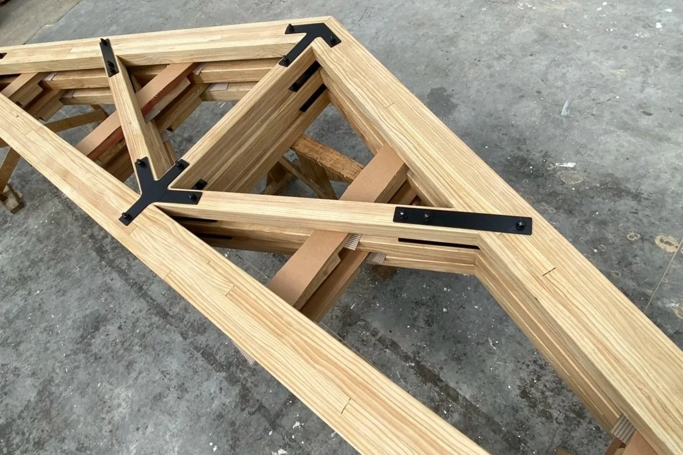 A wooden frame structure with black metal brackets, placed on a workbench in a woodworking shop.