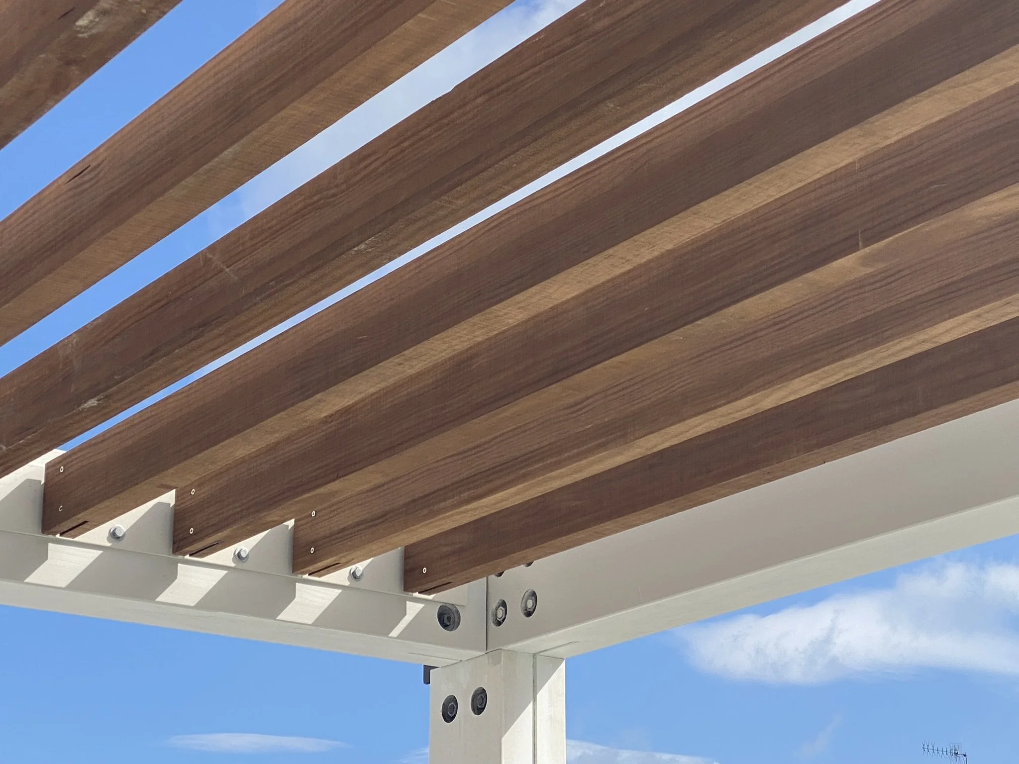 Close-up of a wooden pergola with white beams and bolts, against a blue sky with some clouds.