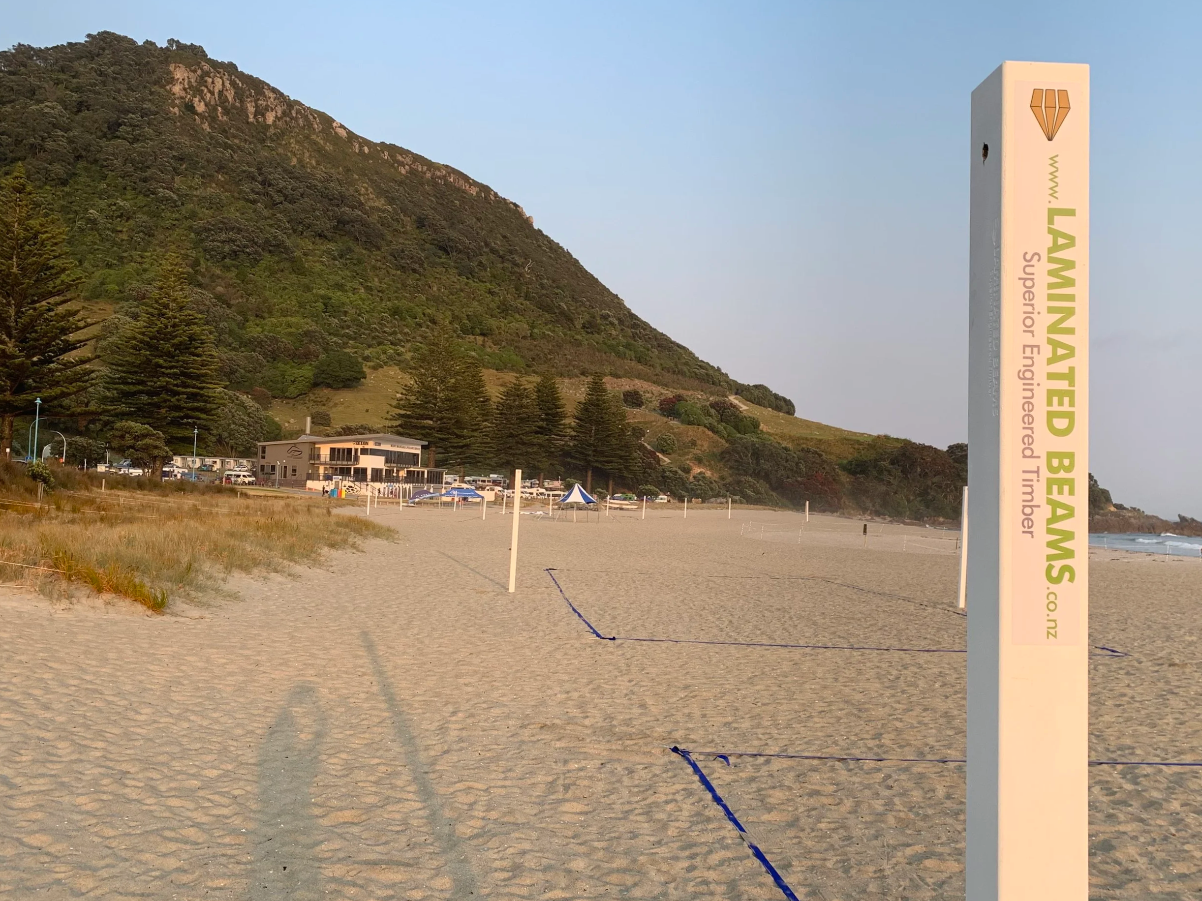 A sandy beach with a coastline, a large mountain covered with greenery in the background, and a signpost on the right side reading 'Laminated Bed' with a diamond-shaped logo above it. There are some buildings and tents near the beach and several trees along the land.