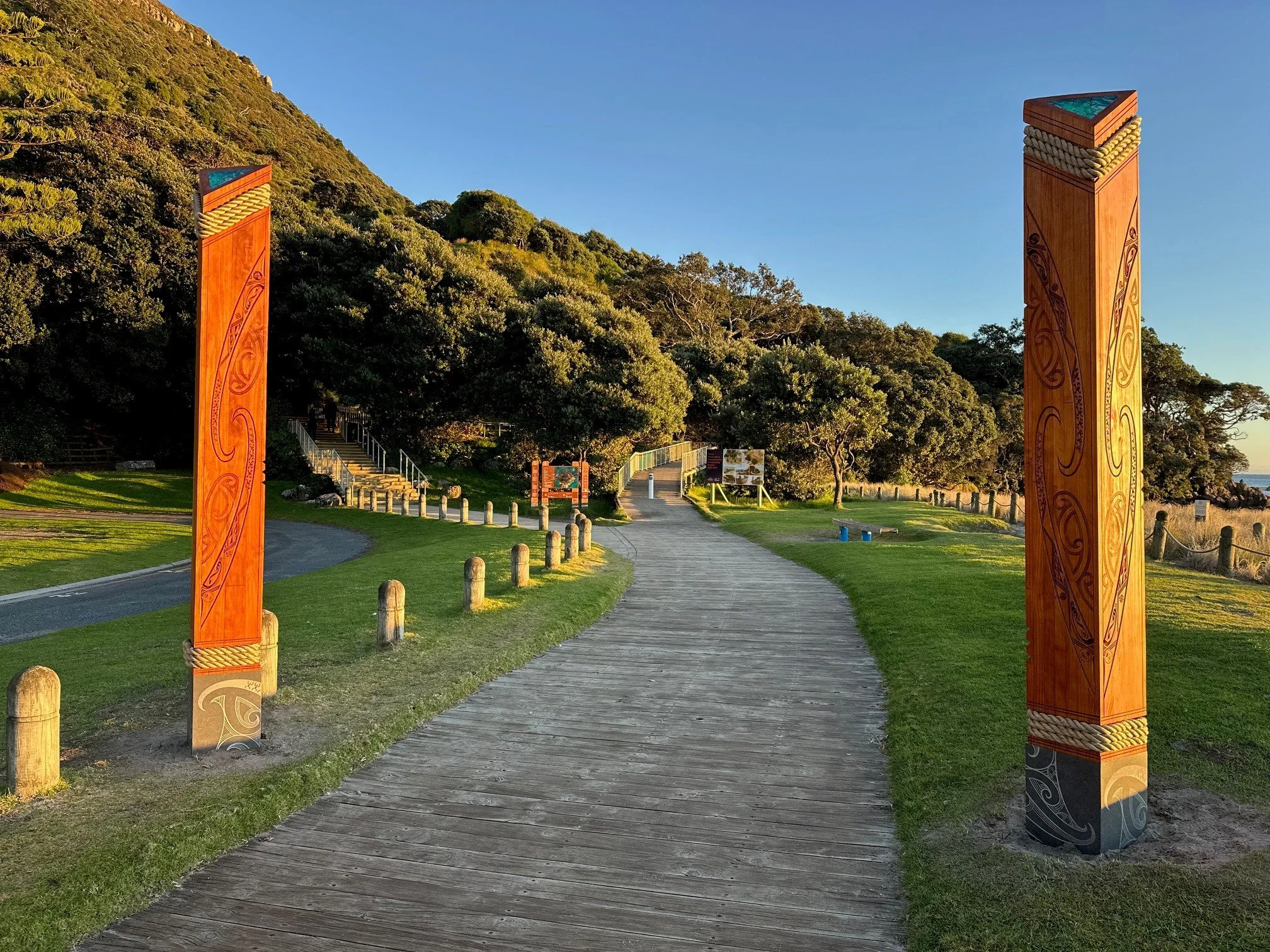 Pathway leading into a park with two tall, carved wooden totem poles on either side. There are stairs and informational signs further along the path, surrounded by trees and a hillside, with the sky clear and sunny.