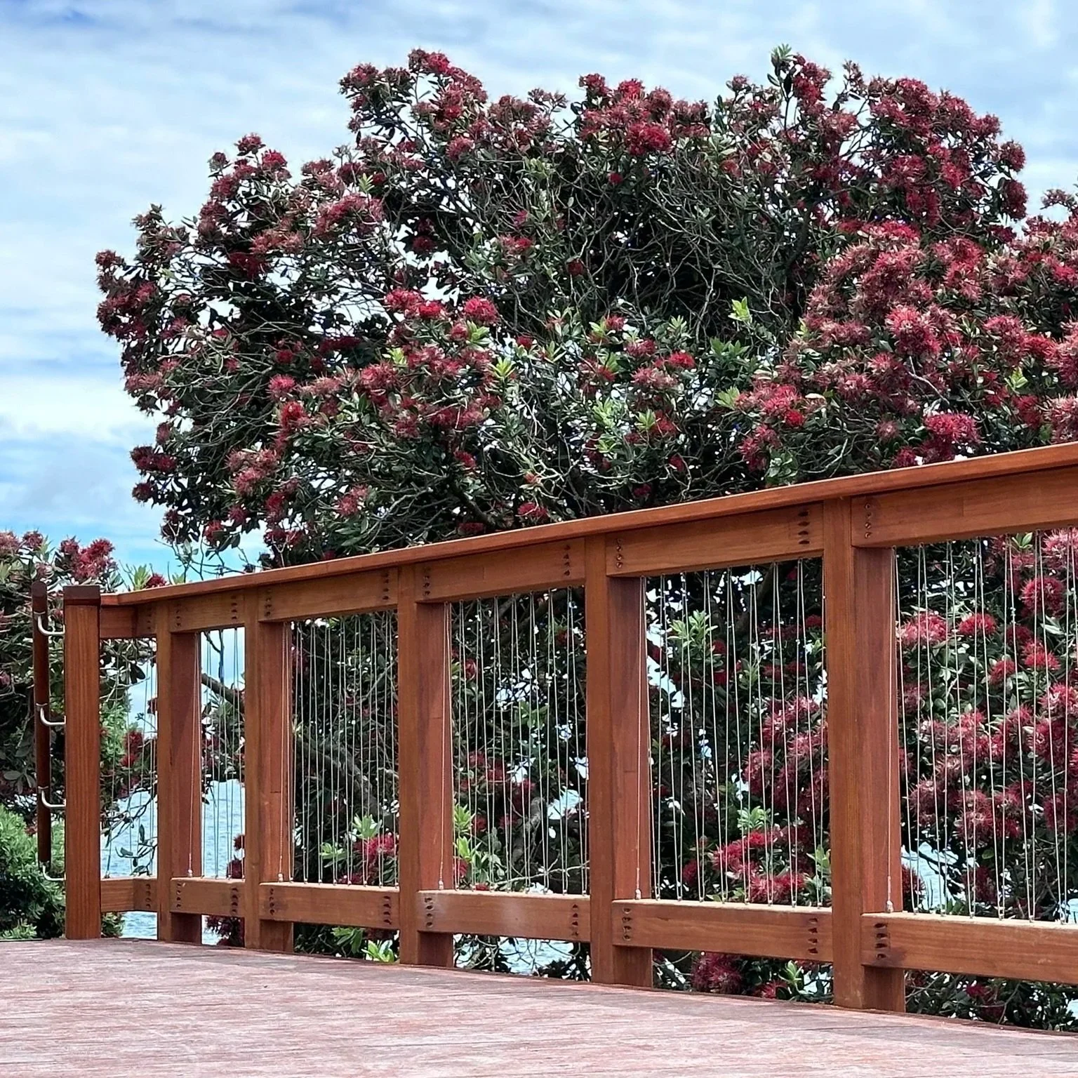 A wooden deck with a railing in the foreground and a large shrub with pink flowers in the background under a cloudy sky.