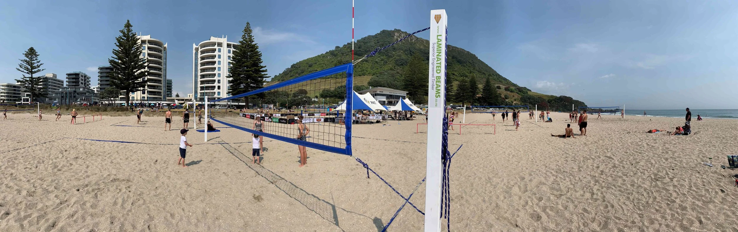 Beach scene with people playing beach volleyball, sand, ocean, and buildings in the background.