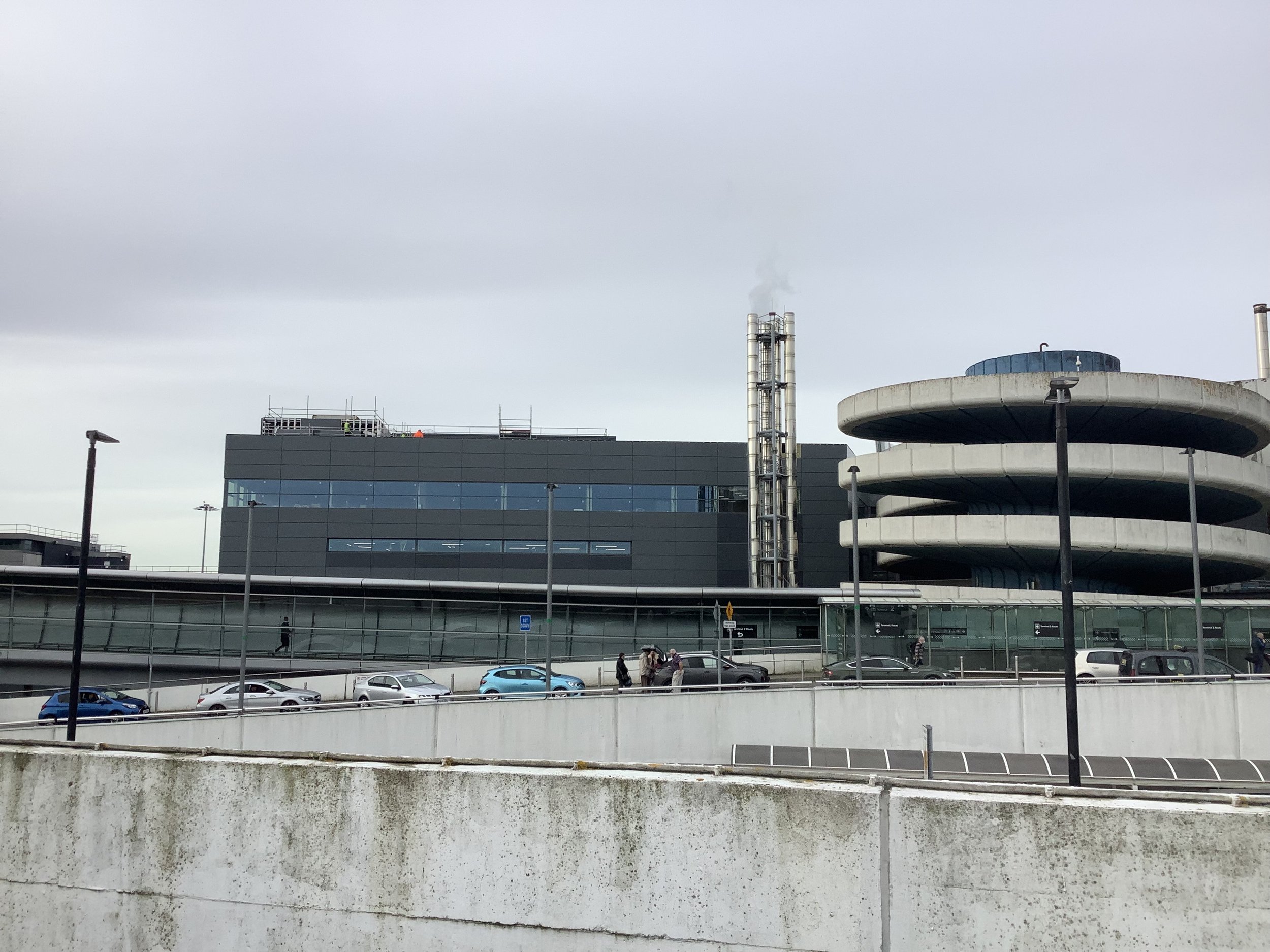 Modern airport terminal building with cars parked outside and overcast sky.