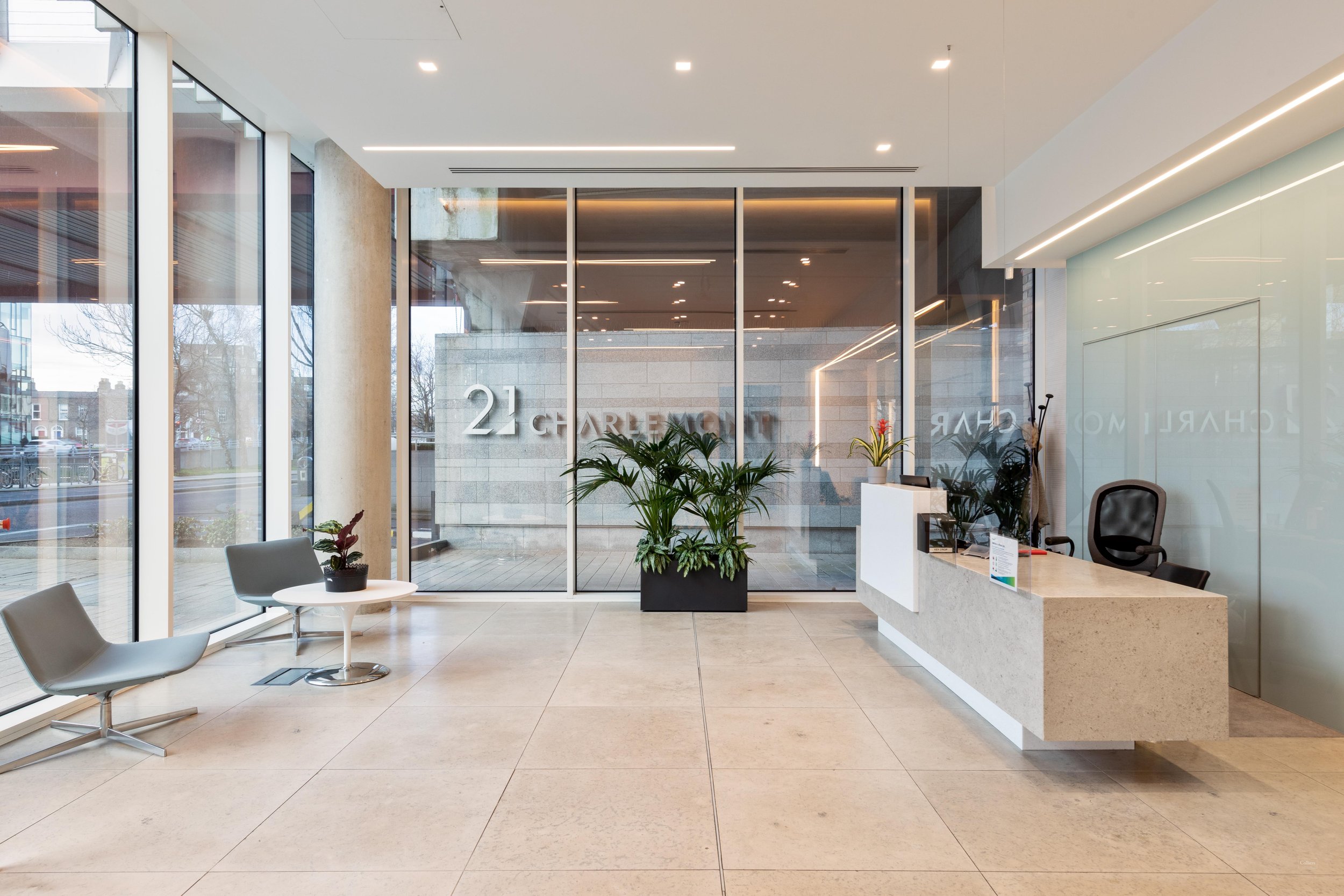 Lobby of a modern building with large glass windows, a reception desk on the right, a few chairs and a small table on the left, and a large potted plant in the center. The building's address, 21 CHARLESTON, is visible on the glass.