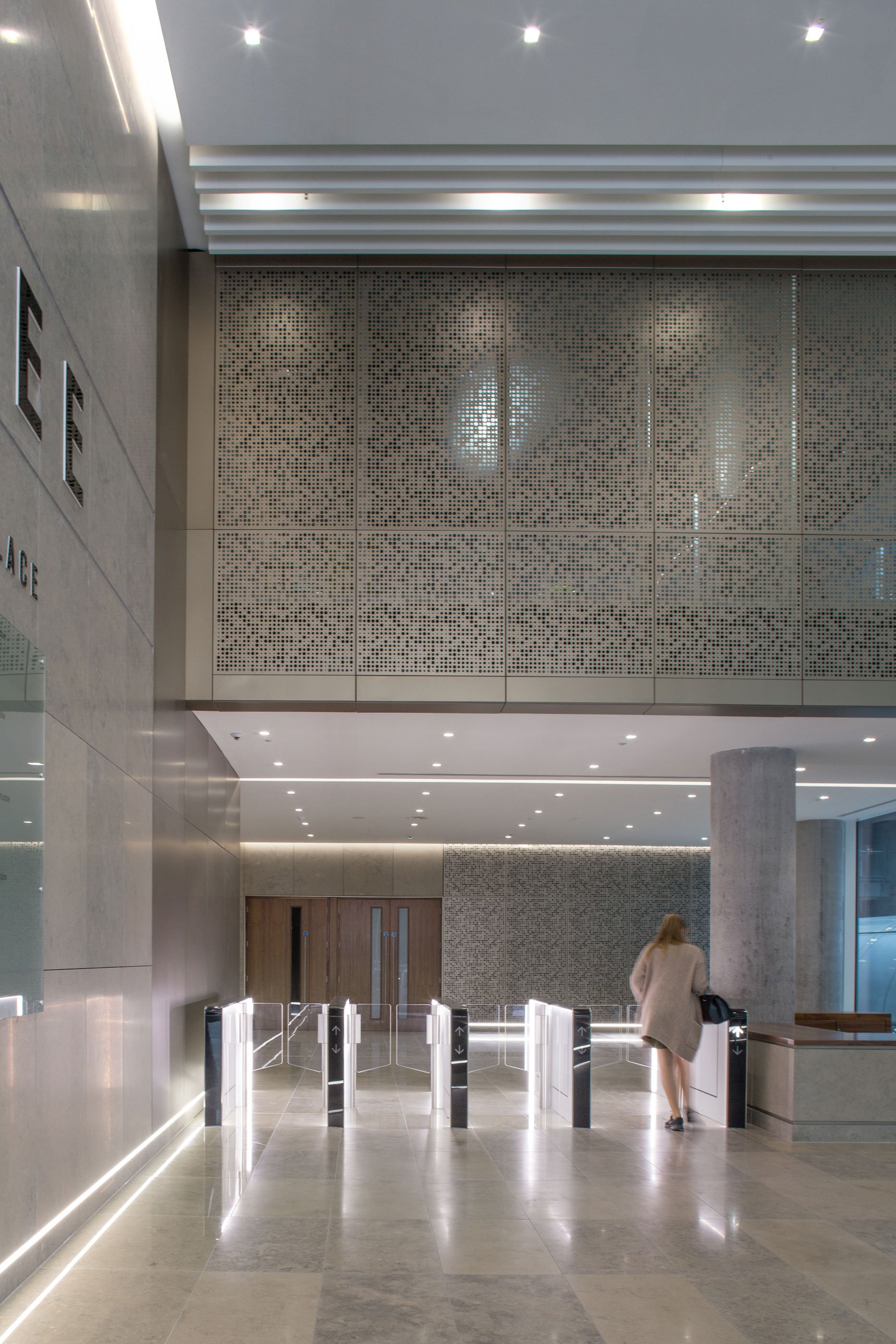 Interior view of a modern building lobby with a woman walking toward turnstiles, polished floors, a large column, and ceiling with recessed lighting.