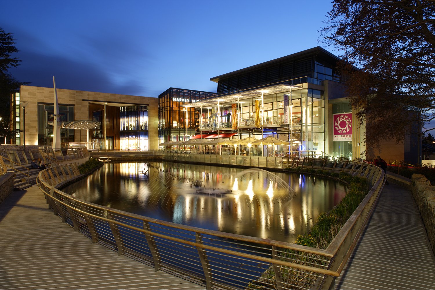 Night view of a modern shopping mall or entertainment complex with glass facades, lit up interiors, and a reflecting pool in front. There are outdoor seating areas with umbrellas and a winding wooden walkway along the water.