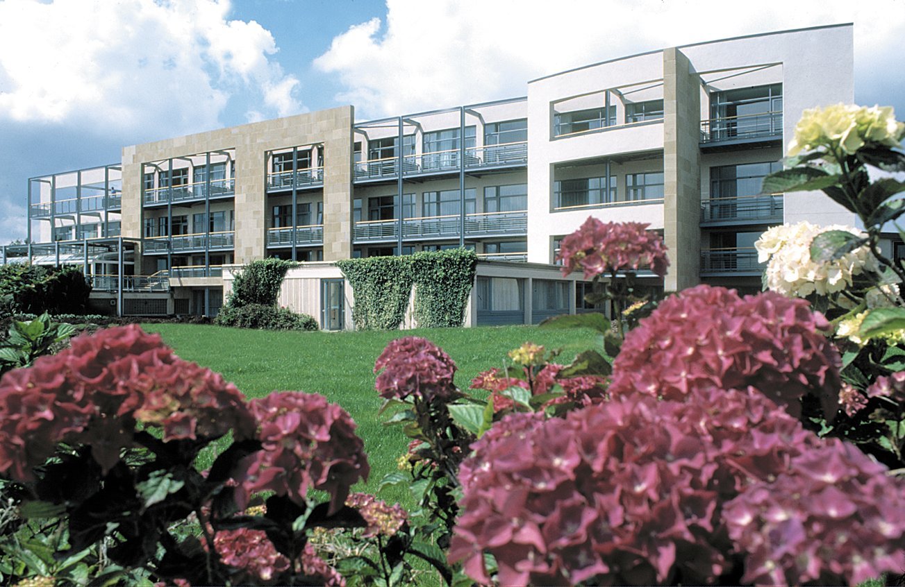 Modern multi-story apartment building with balconies, surrounded by a green lawn and colorful hydrangea flowers in the foreground, under a partly cloudy sky.