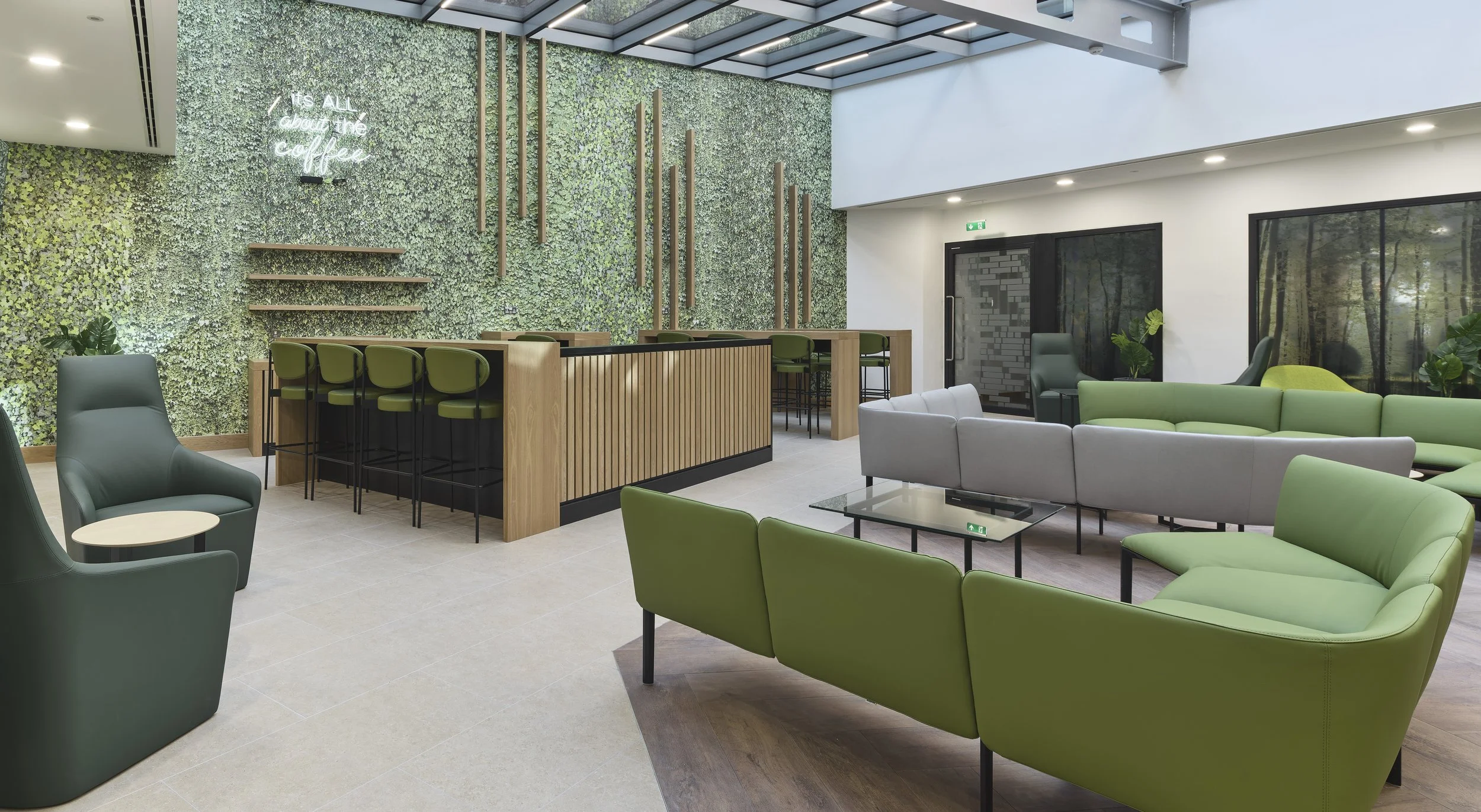 Modern coffee shop with green and gray seating, a green living wall, bar area with green chairs, indoor plants, and large windows showing trees outside.