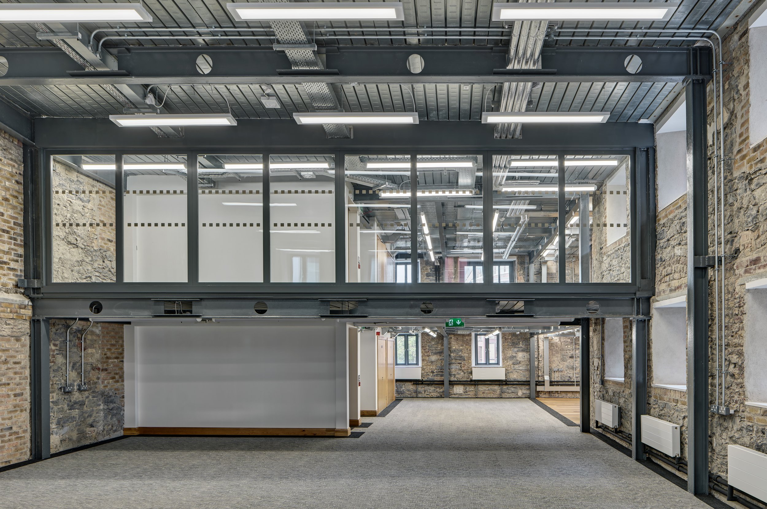 Empty office space with exposed brick walls, carpeted floors, and a glass-enclosed mezzanine level.