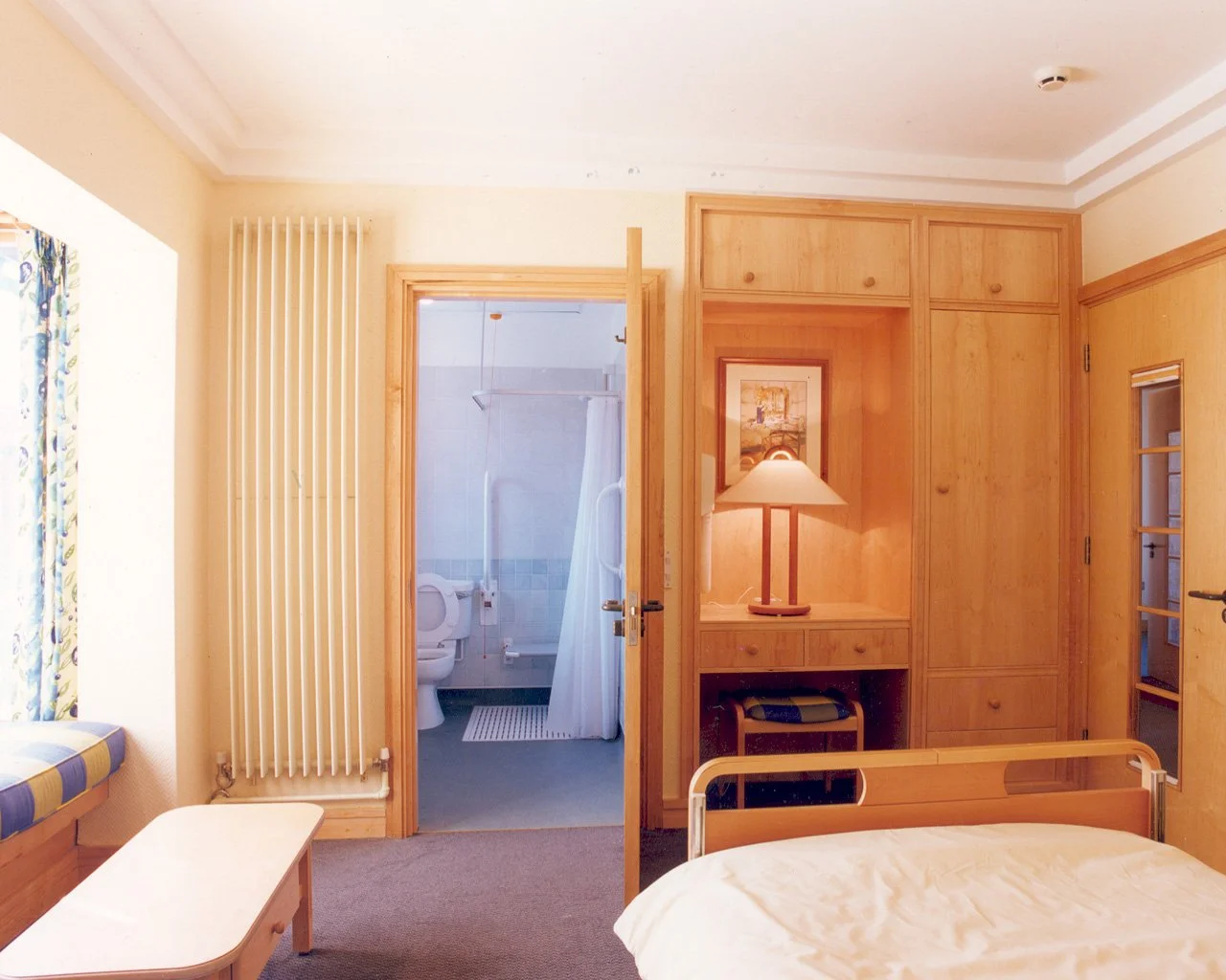 Interior view of a bedroom with a partially visible bed, wooden furniture, a table, a dresser with a lamp, and a bathroom door open showing a toilet and shower.