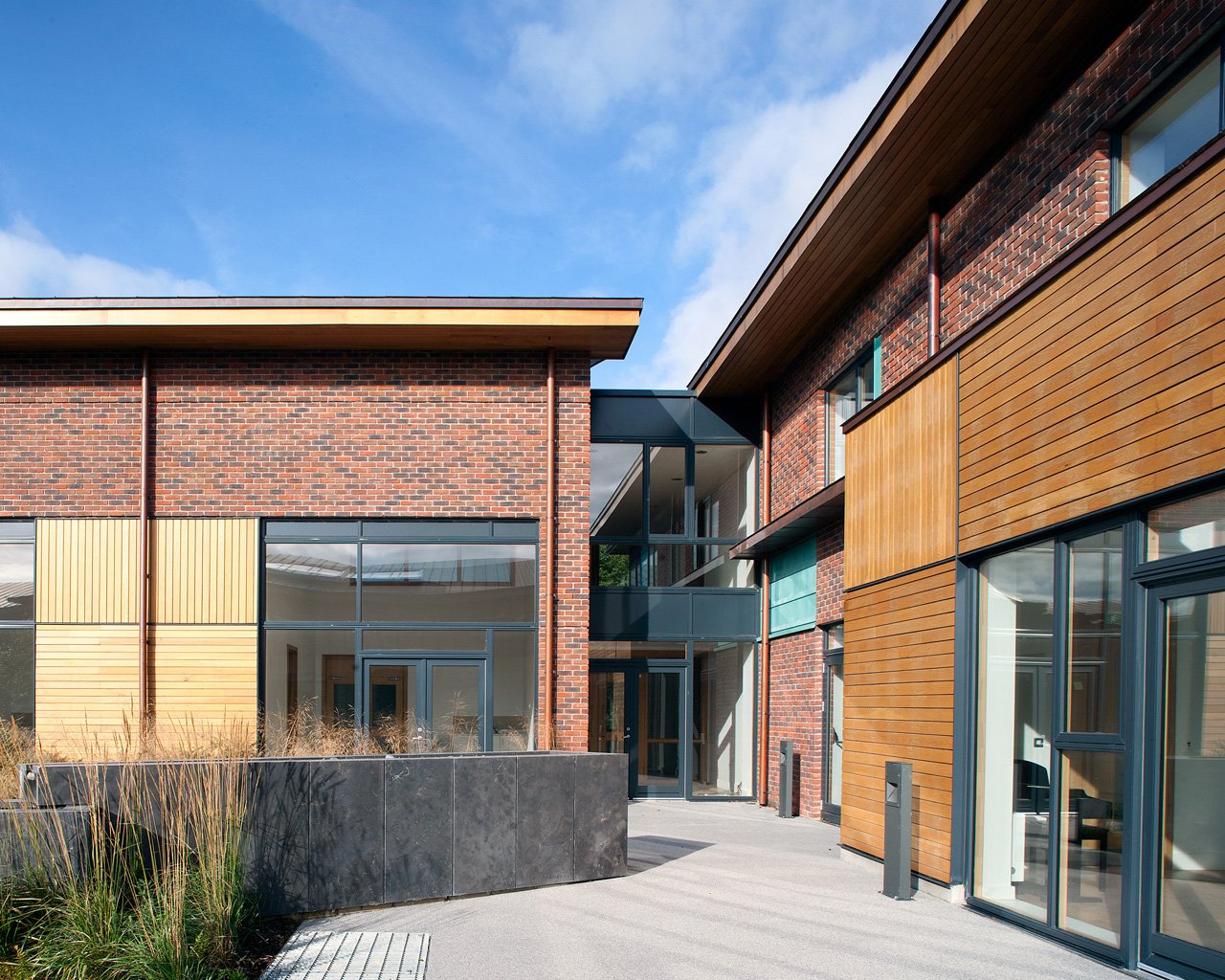 Modern building with brick and wooden exterior, glass windows and entrance, and landscaped area in front under a blue sky.