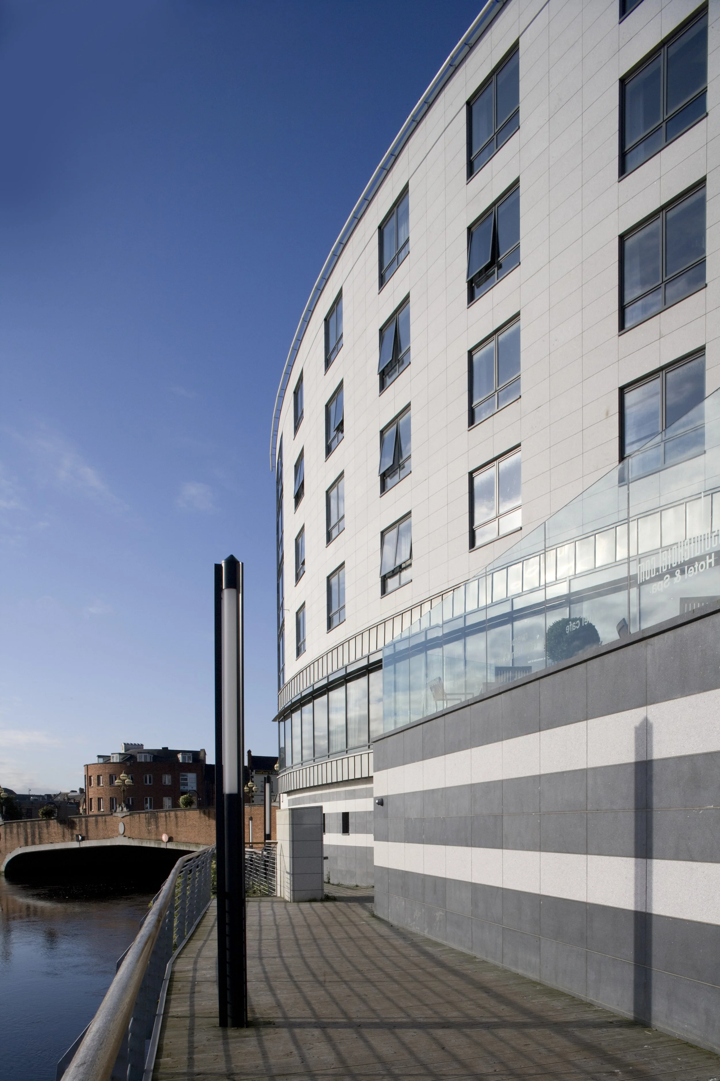 Modern multi-story building by a waterway with a walkway and railing, reflective glass windows, clear blue sky