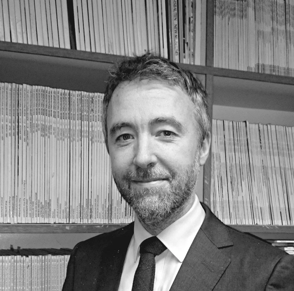 Black and white photo of a man in a suit and tie, smiling, standing in front of a bookshelf filled with magazines or books.