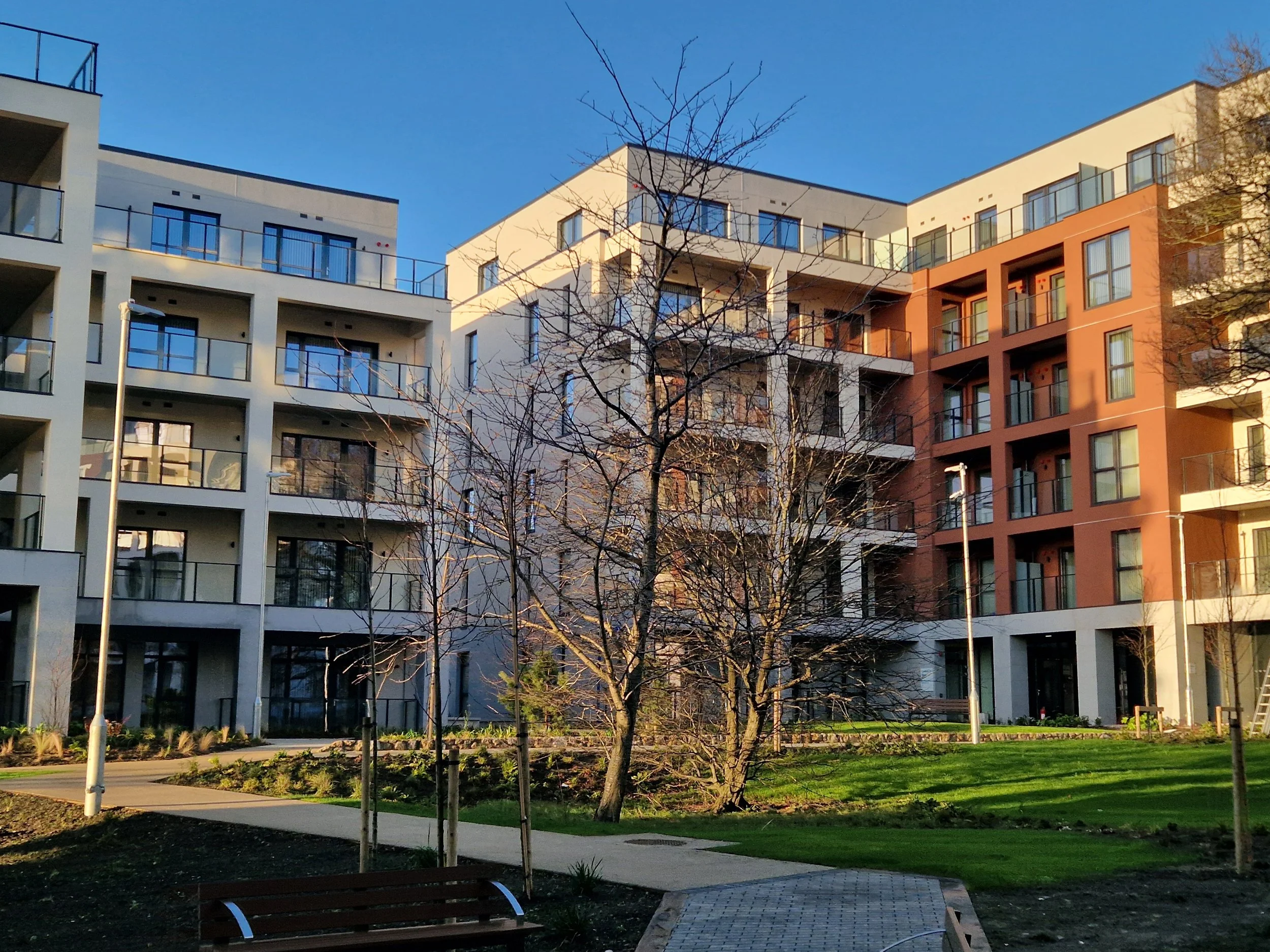 Photograph of modern multi-story apartment buildings with glass balconies, a landscaped courtyard with a path, benches, and leafless trees, under a clear blue sky.