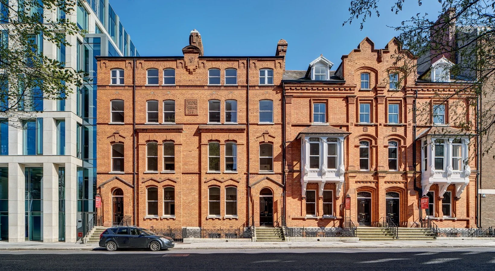 A row of historic brick townhouses with detailed white bay windows on a city street, adjacent to a modern glass building, under a clear blue sky.