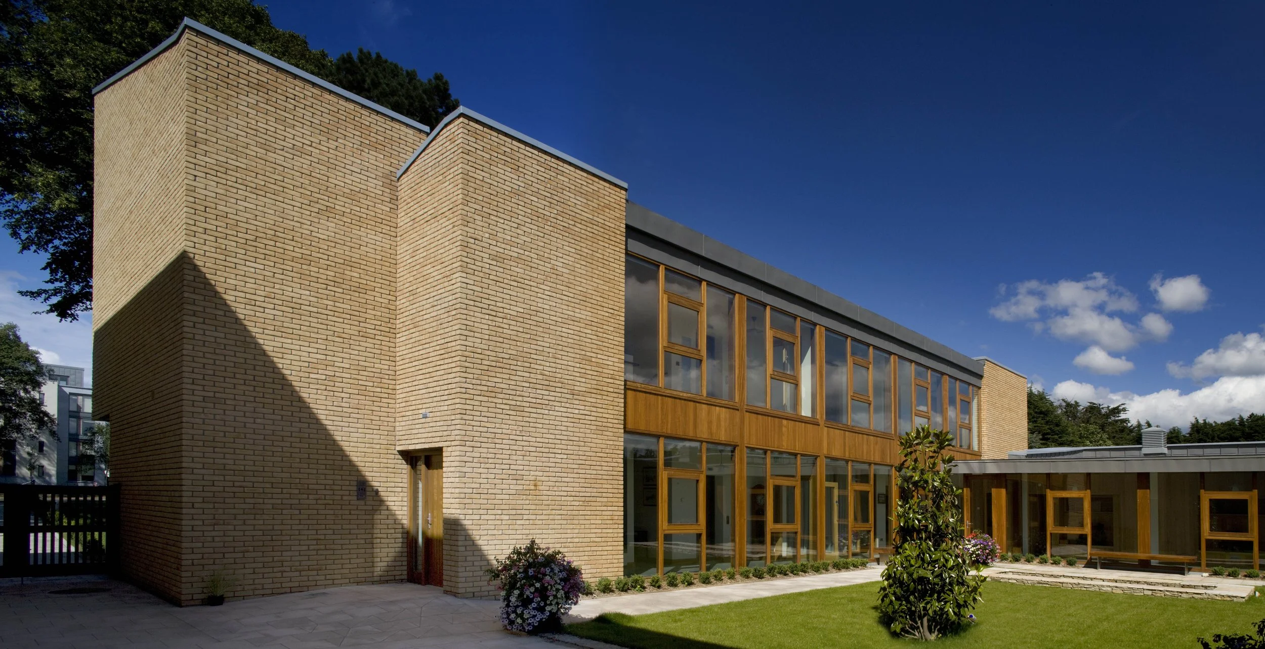 Modern building with brick walls and large wooden-framed glass windows, surrounded by a well-maintained lawn and a blue sky with some clouds.