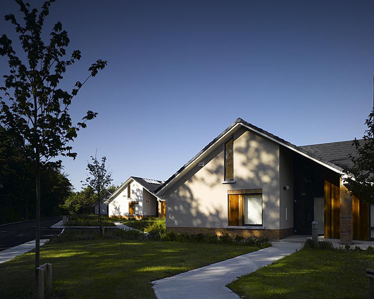Modern suburban houses with white walls, wooden accents, and sloped roofs, surrounded by green lawn and trees, during dusk.