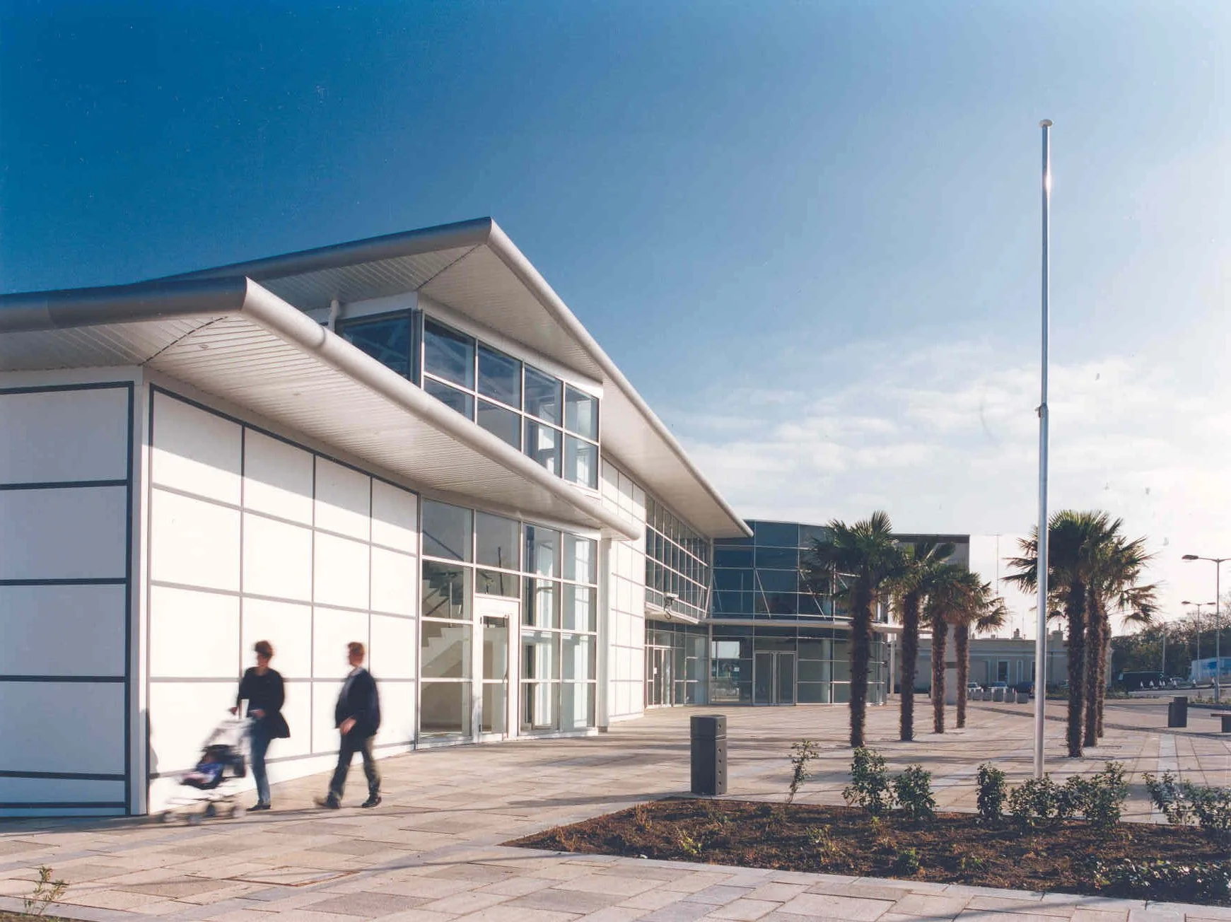 Modern glass building with palm trees outside and two people walking with a stroller in front, under a clear sky.
