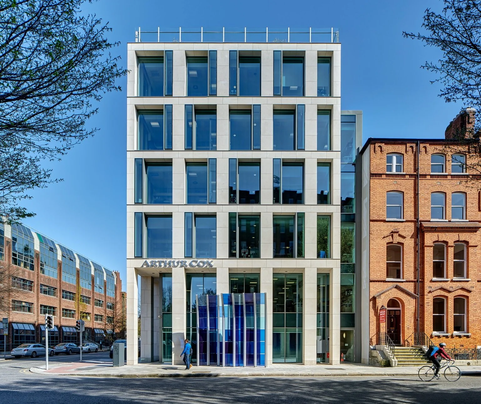 Modern white and glass office building with the sign 'Arthur Cox' on the front. Blue sky and trees on either side, with a person walking and a cyclist passing by on the street in front.