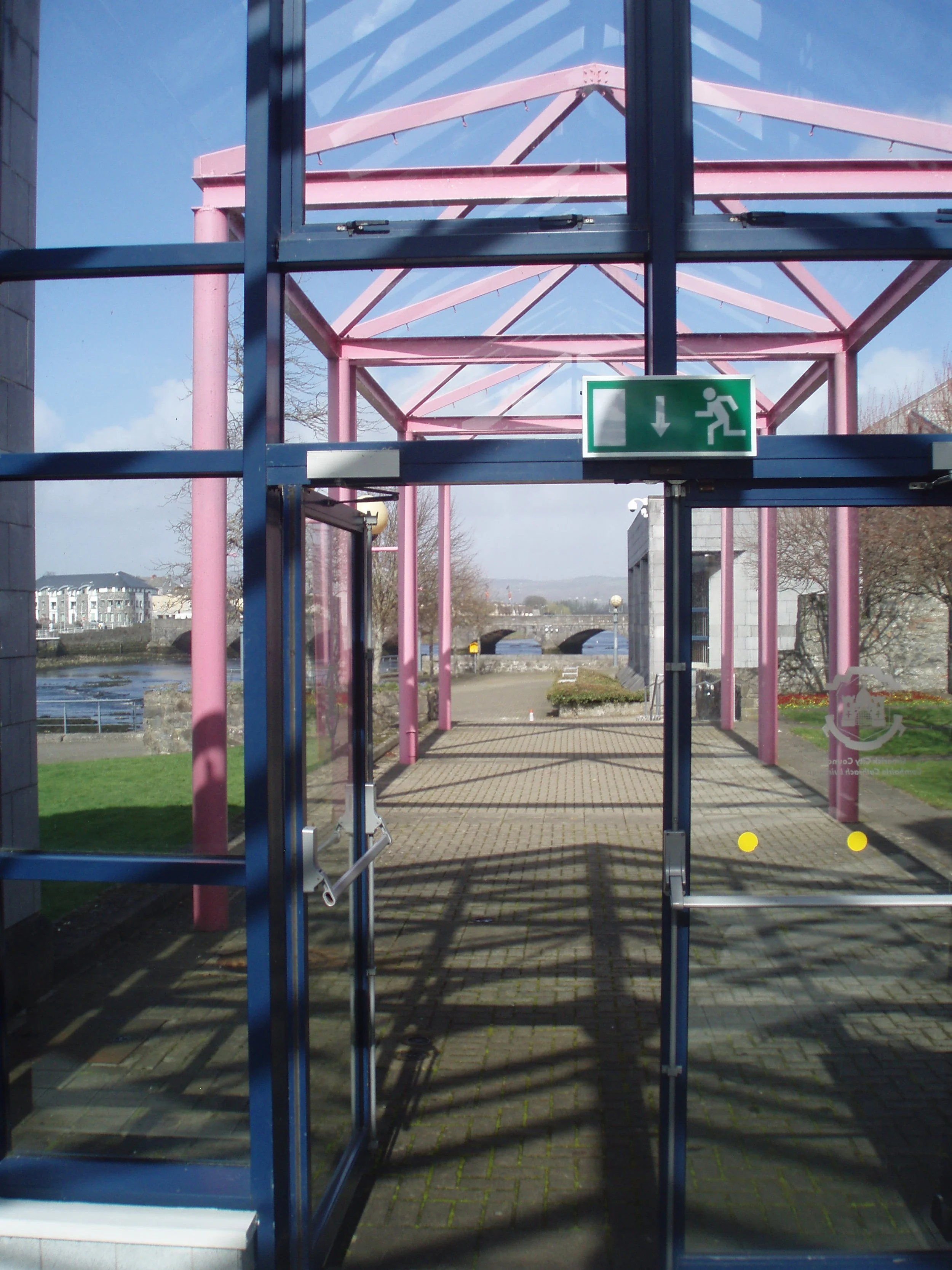 View through glass doors of pink and blue metal structure in an outdoor park area with a river, bridge, and buildings in the background.