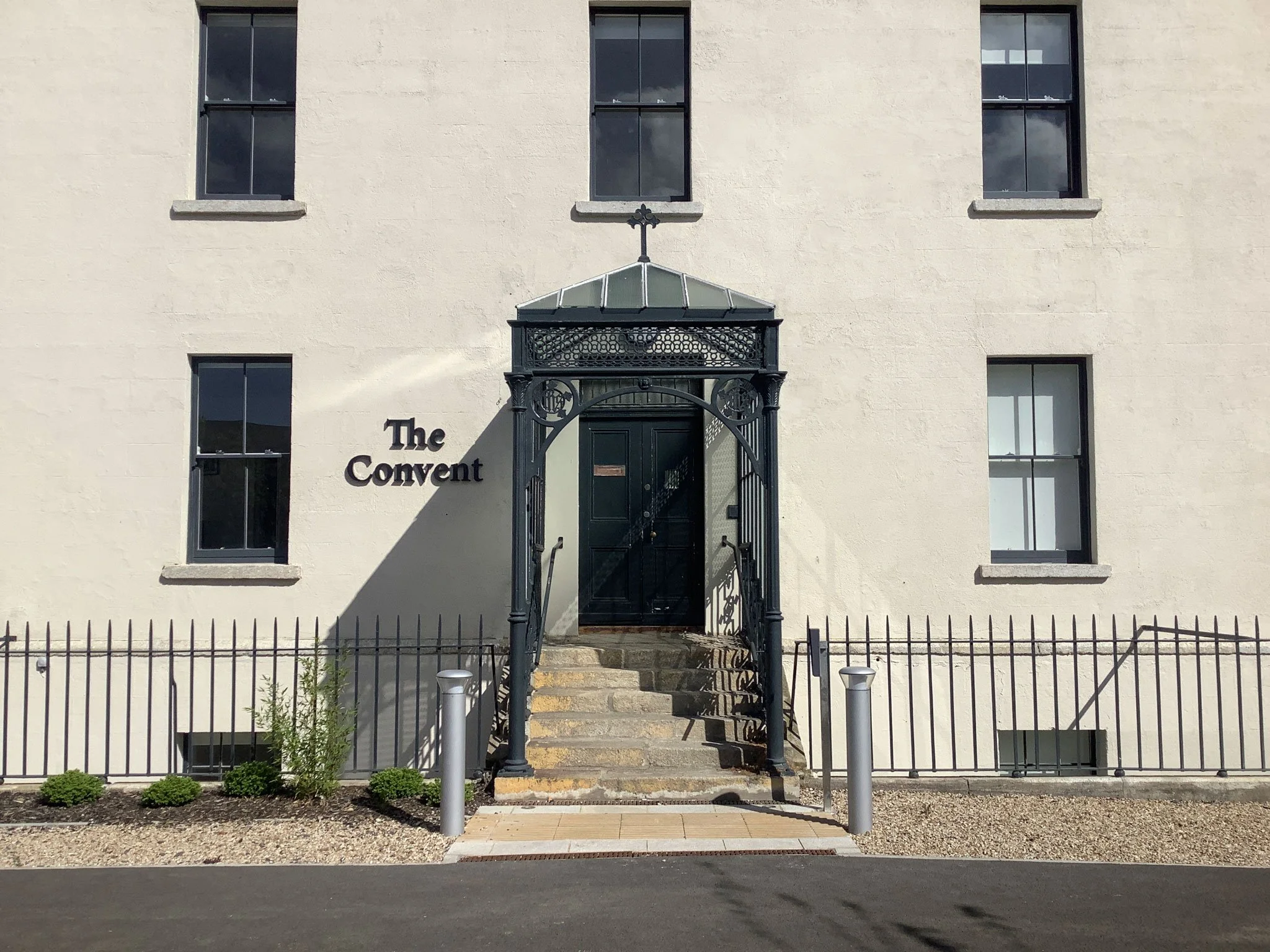 Exterior of a white building labeled 'The Convent' with a black door and an ornate iron canopy over the entrance, stairs leading up to the door, and a small garden with bushes and a fence.