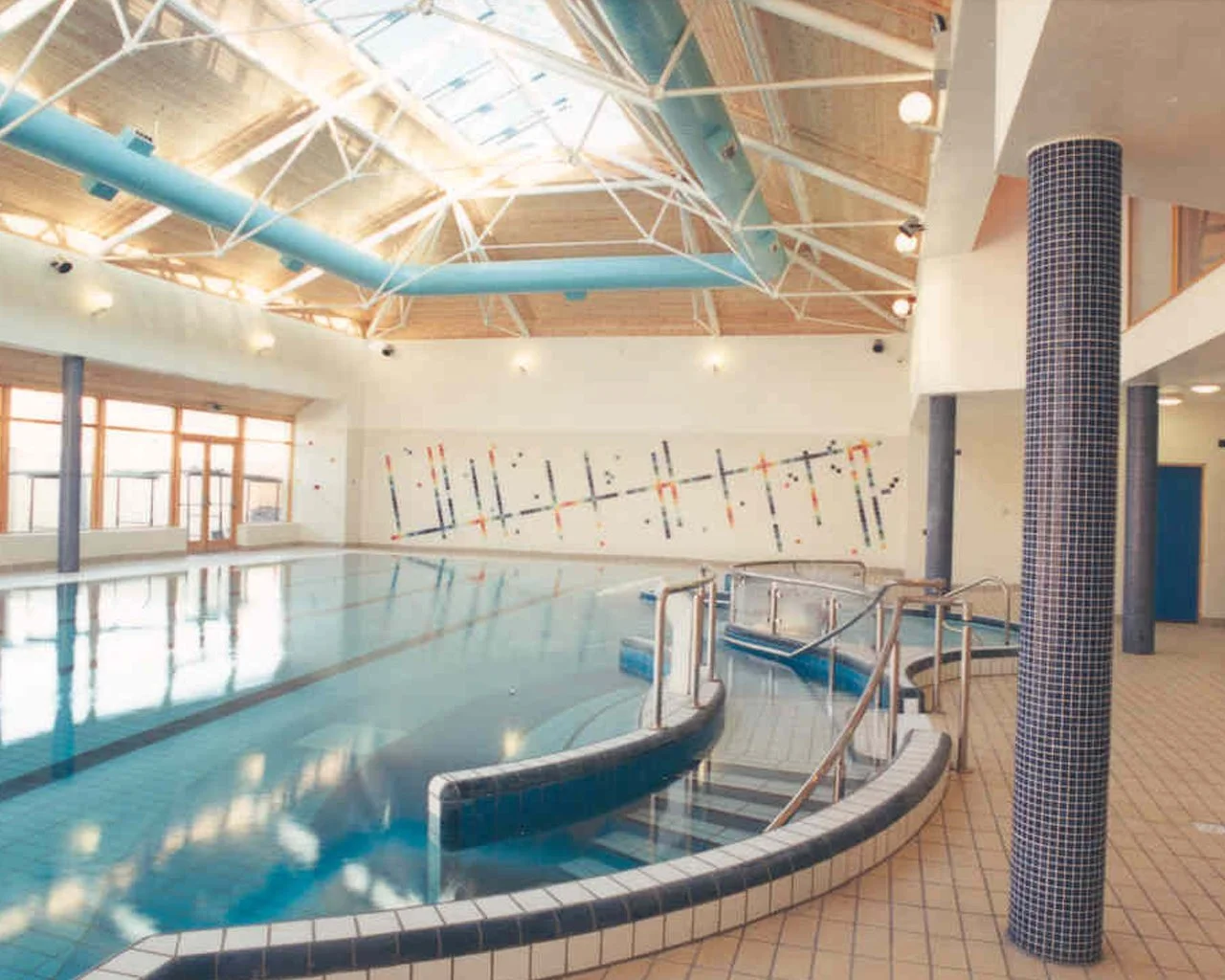 Indoor swimming pool area with a tiled pool, stainless steel handrails, and a decorative wall mural. Wooden ceiling with skylights and large windows on one side.