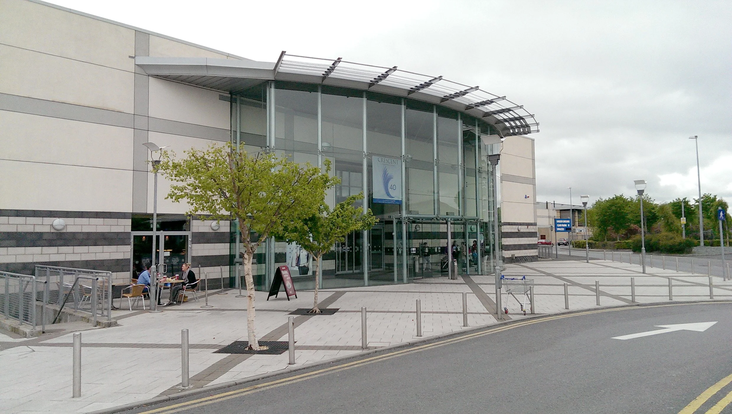 Exterior of a modern shopping mall with glass entrance, trees, and a few people sitting outside at tables.