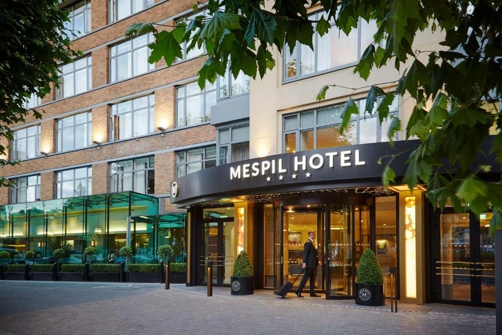 The entrance of the Mespil Hotel with a man walking in front, surrounded by trees and greenery, with a brick hotel building in the background.