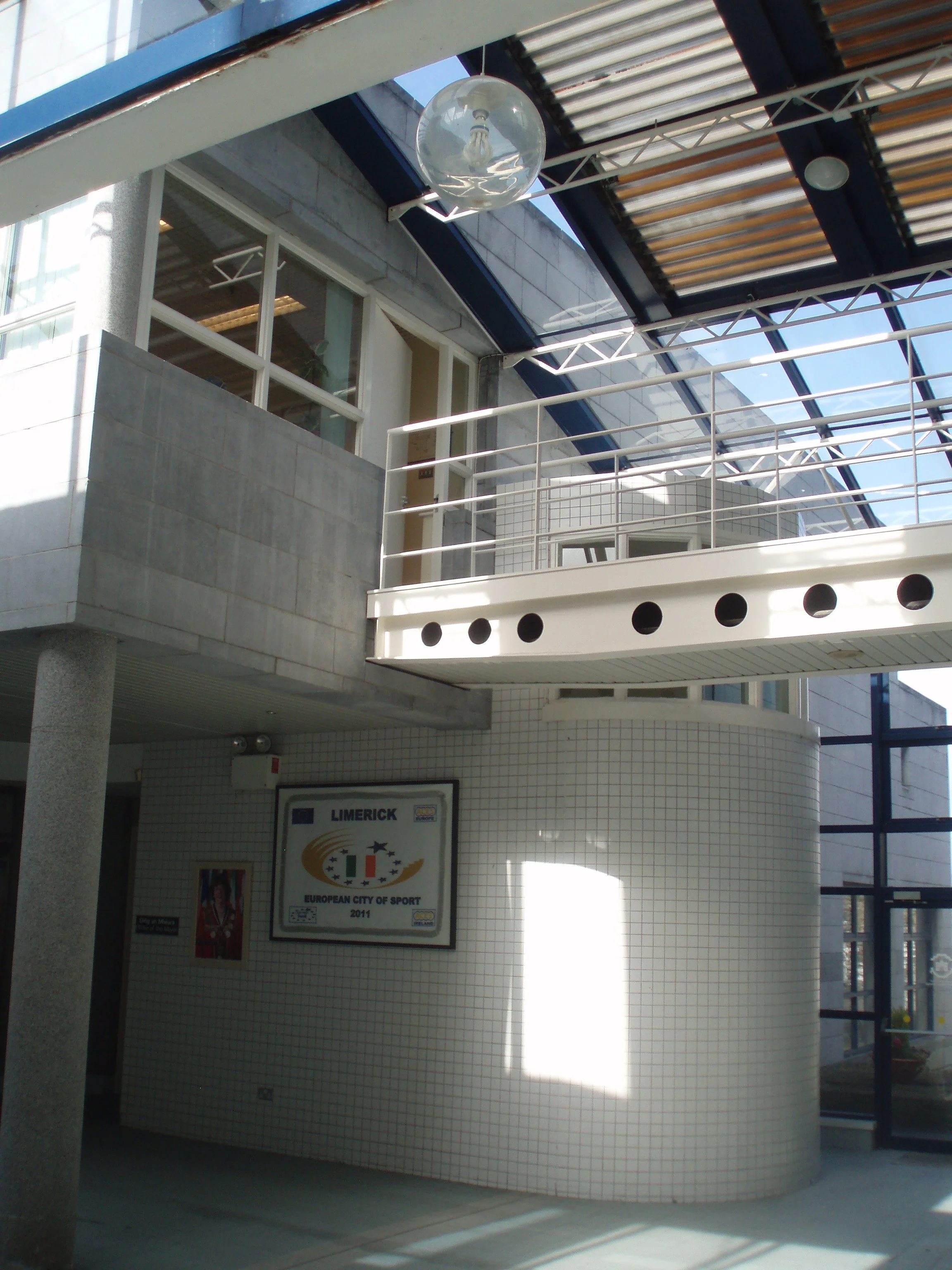 Interior view of a modern indoor sports complex with tiled walls, a sign that reads "Limerick European City of Sport 2011," and an outdoor area with glass and metal structures.