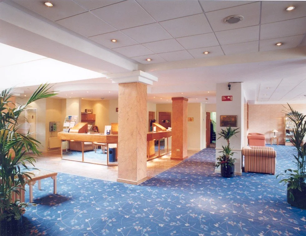 Interior of a hotel lobby with seating areas, potted plants, and a reception desk, with a blue carpet and cream-colored walls.